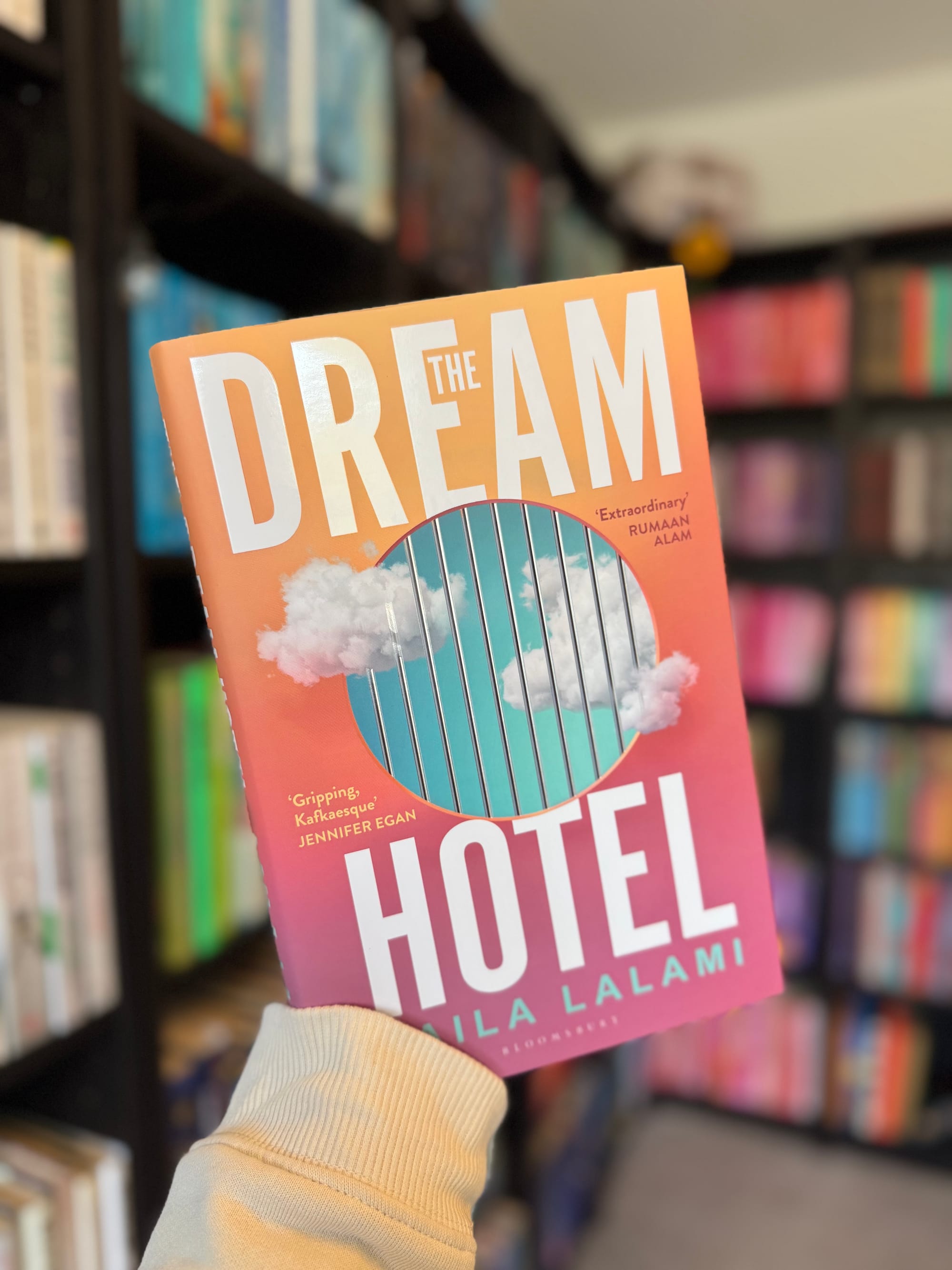 A hand holds "The Dream Hotel" book in a library. The cover features clouds and vertical bars, with an orange to pink gradient background.