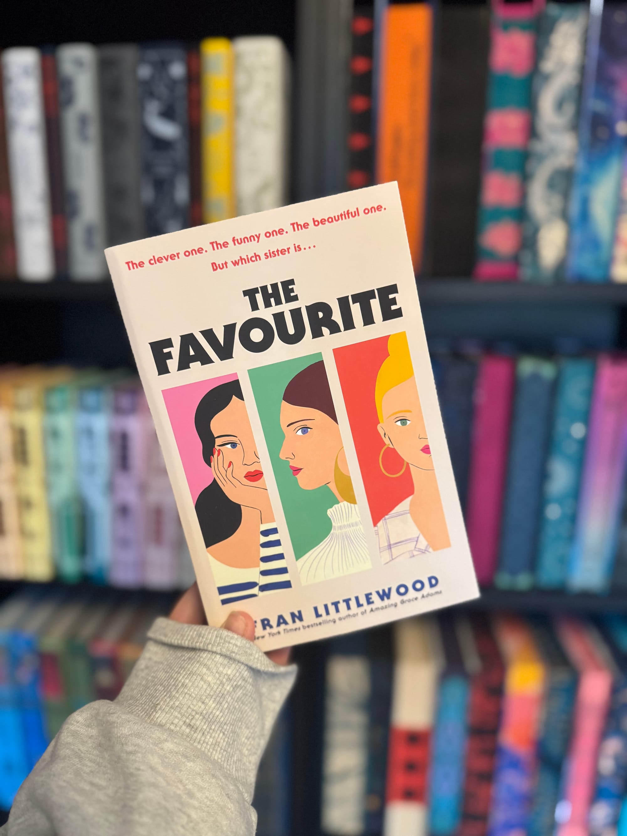 A hand holds a book titled "The Favourite" by Fran Littlewood. The cover shows three illustrated women's faces. Bookshelves are blurred behind.