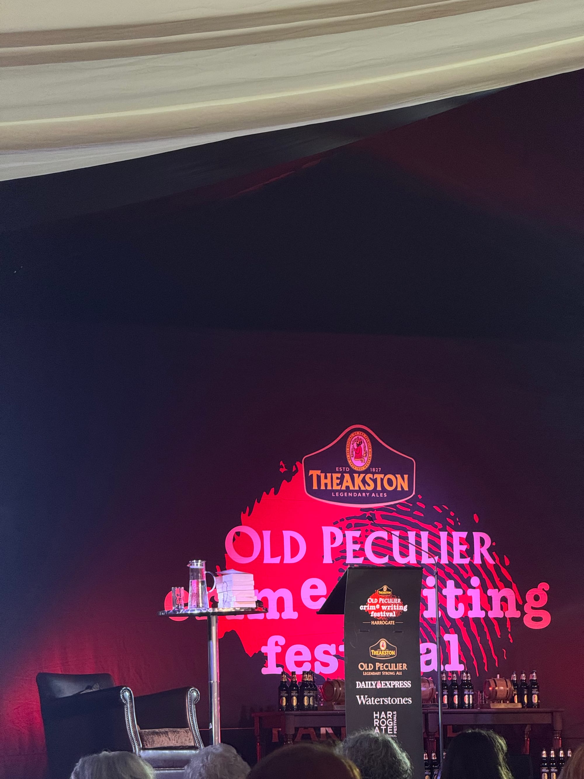 View of the main festival stage set for a panel event, featuring the Theakston Old Peculier Crime Writing Festival logo, podium, two chairs, and stacks of books against a dramatic black and red backdrop.