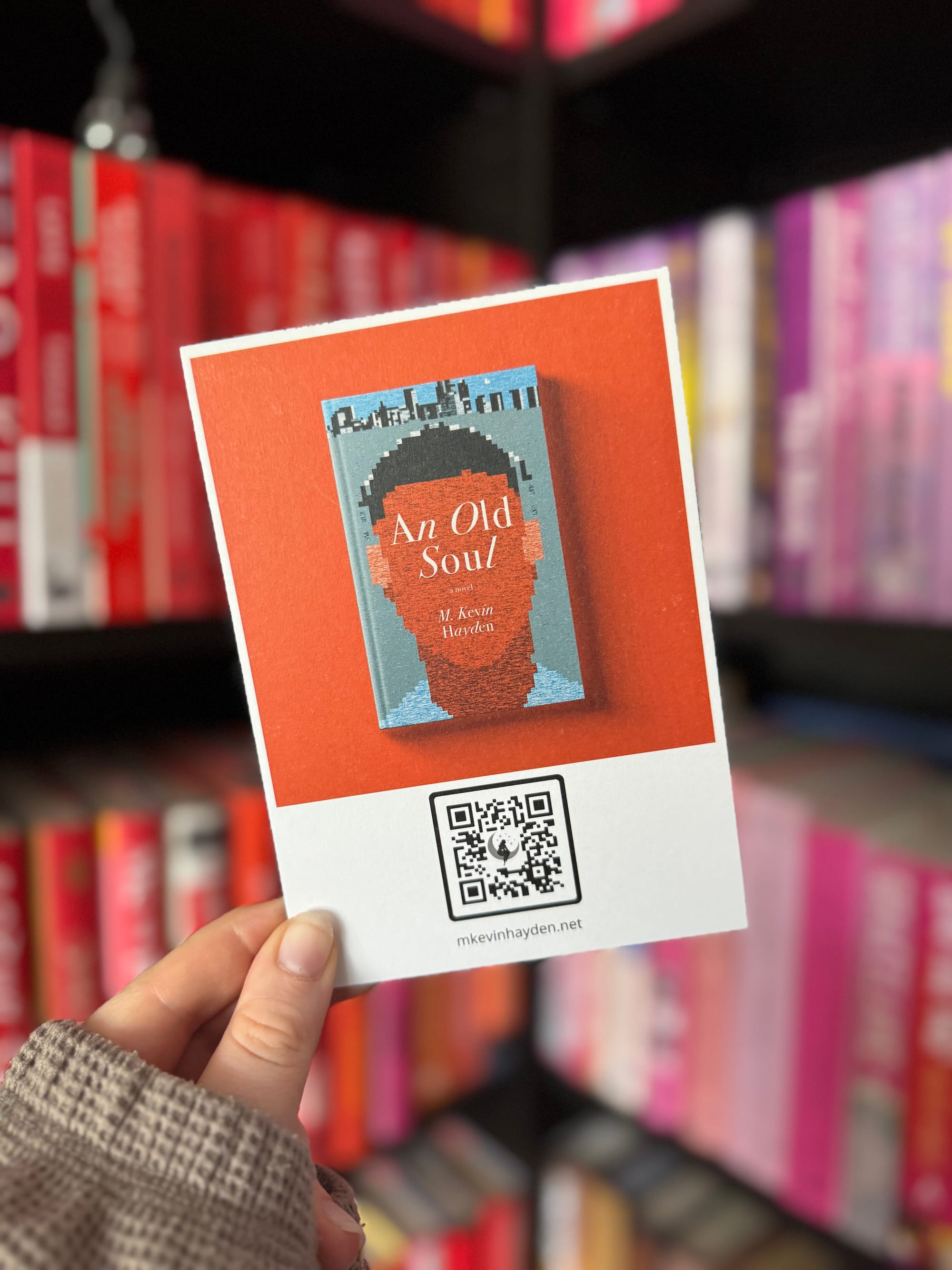 A hand holds a promotional card for the book An Old Soul by M. Kevin Hayden. The card features the book cover, which has a pixelated silhouette on a red-orange background, and a QR code below linking to the author's website (mkevinhayden.net). Colour-coordinated bookshelves with red, pink, and purple spines are blurred in the background.