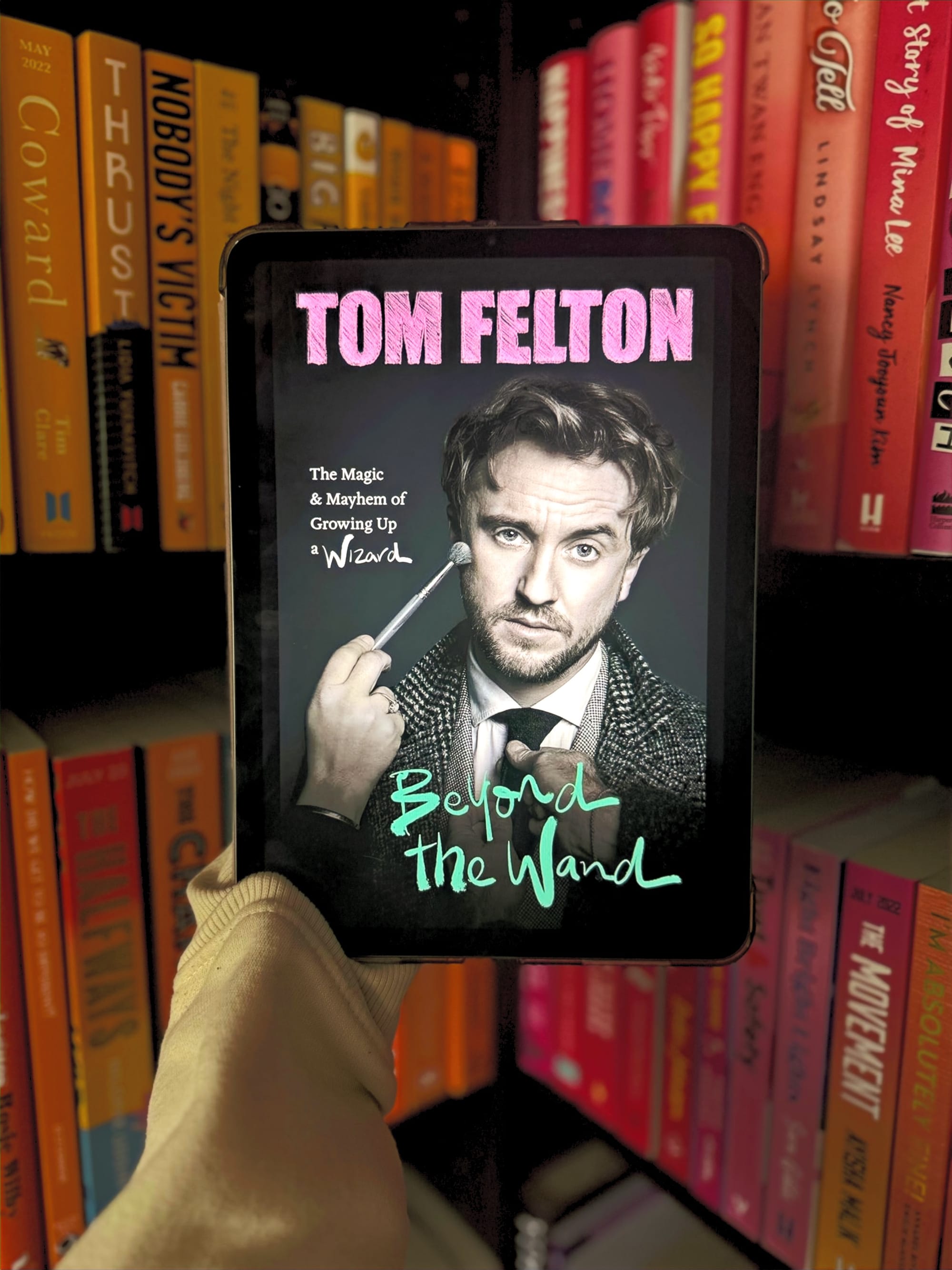 A hand holds a tablet displaying "Tom Felton: Beyond the Wand" book cover. Vibrant bookshelves form the colorful background.