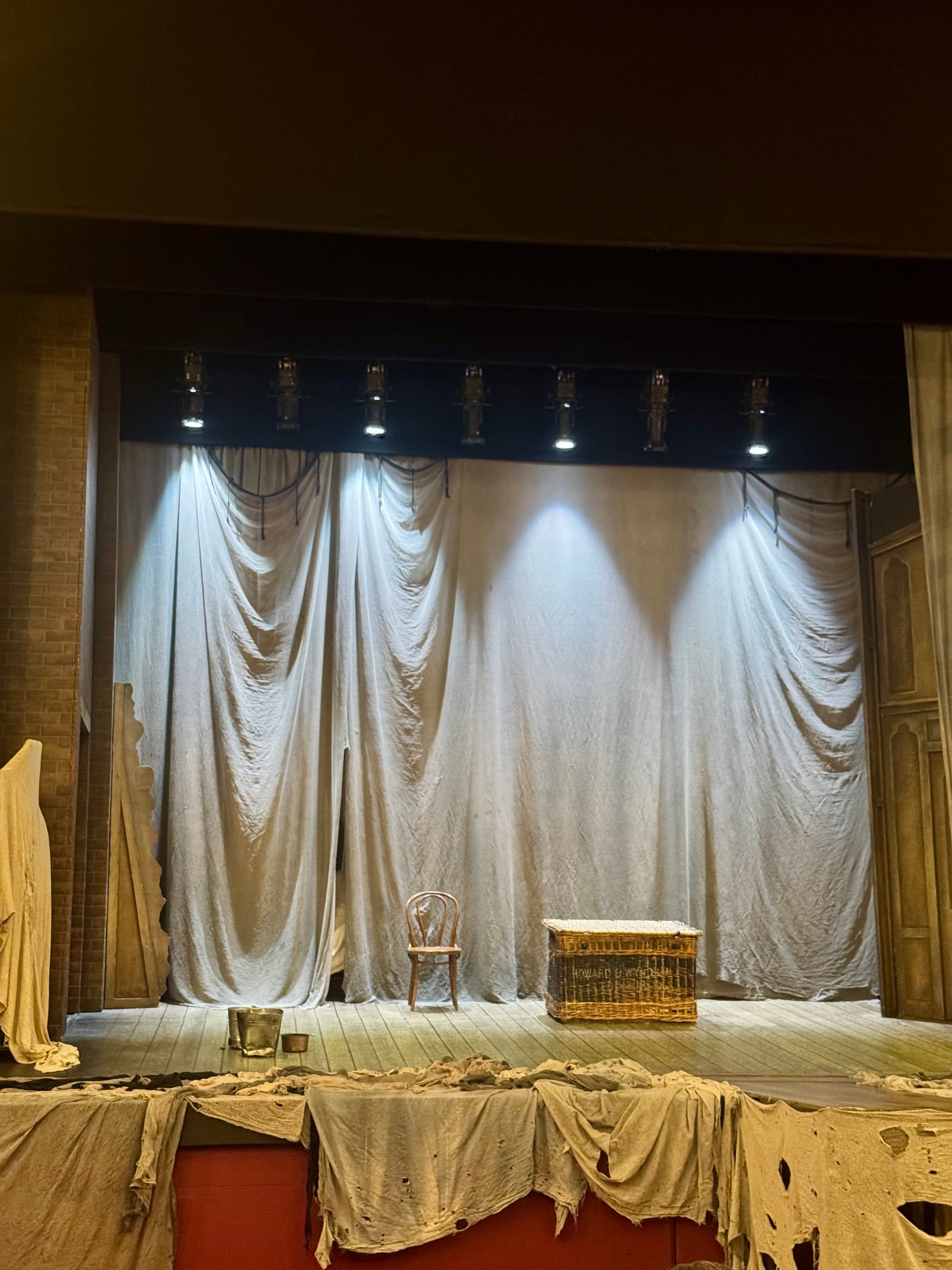 Empty stage with draped curtains, a wooden chair, and a wicker basket labeled "Honage." Dim lighting creates a mysterious atmosphere.