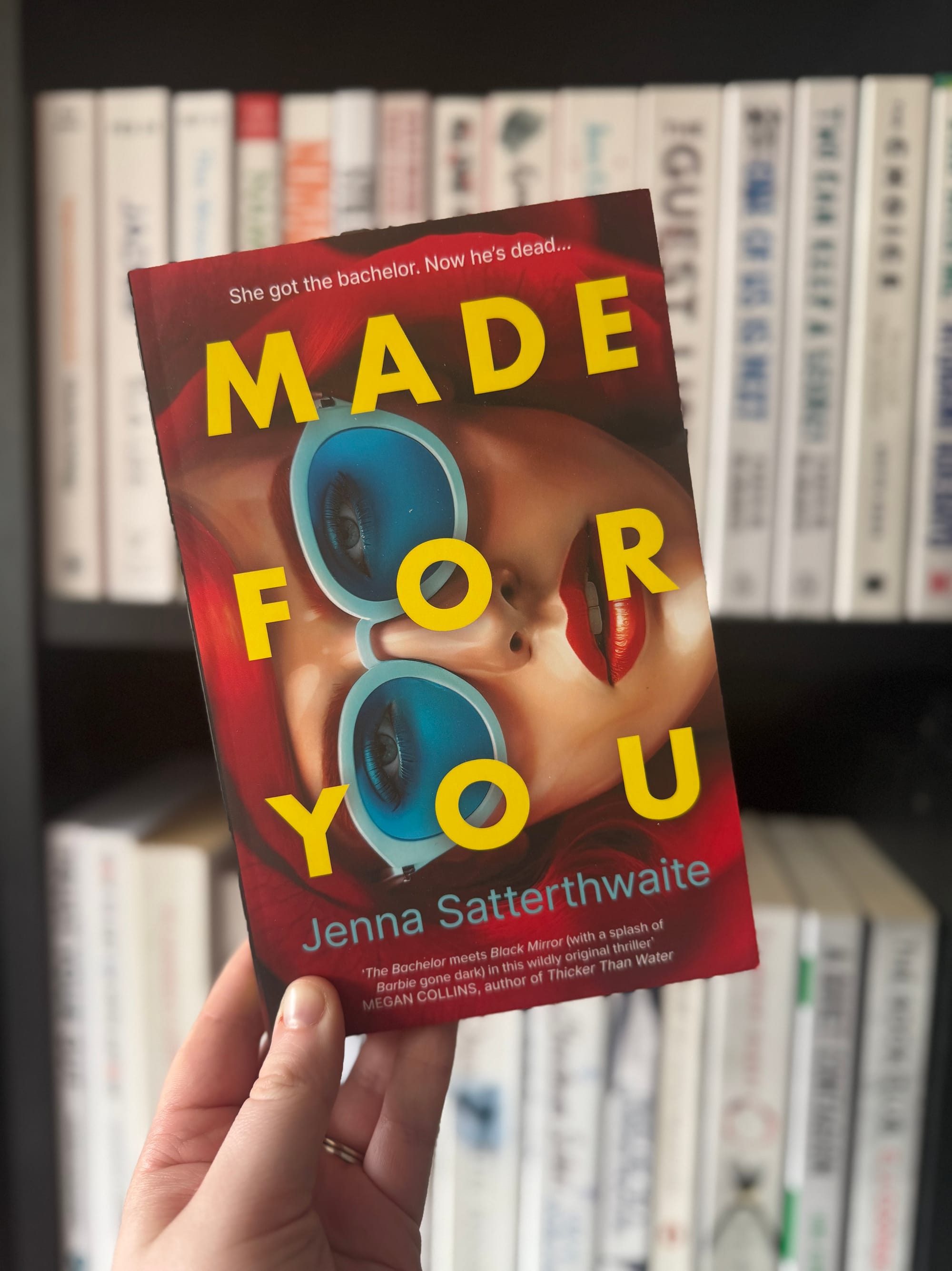 Hand holding the book "Made For You" by Jenna Satterthwaite with vibrant cover art and text. Bookshelf with various books in background.