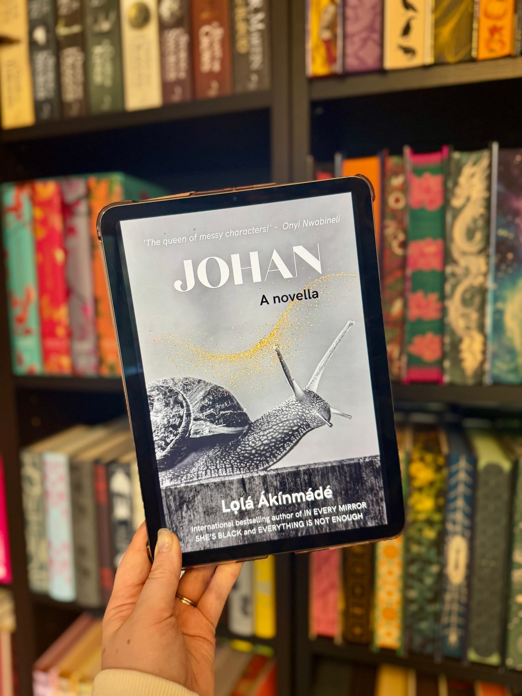 Hand holding a tablet displaying the book cover "JOHAN" by Lola Akinmade, featuring a snail, against a colorful bookshelf background.