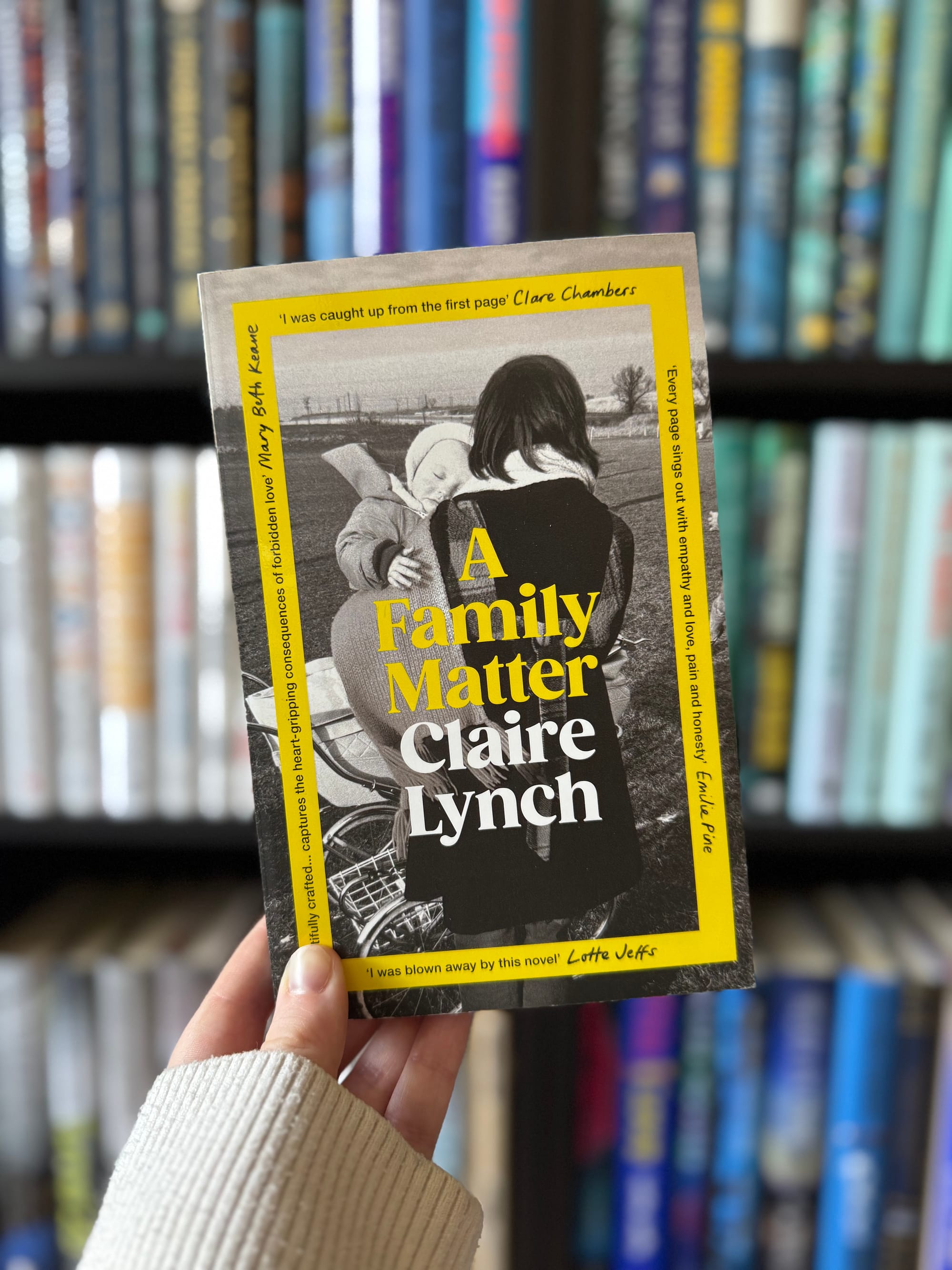 A hand holds a book titled "A Family Matter" by Claire Lynch, with a black-and-white cover of a woman and child. Bookshelf background.