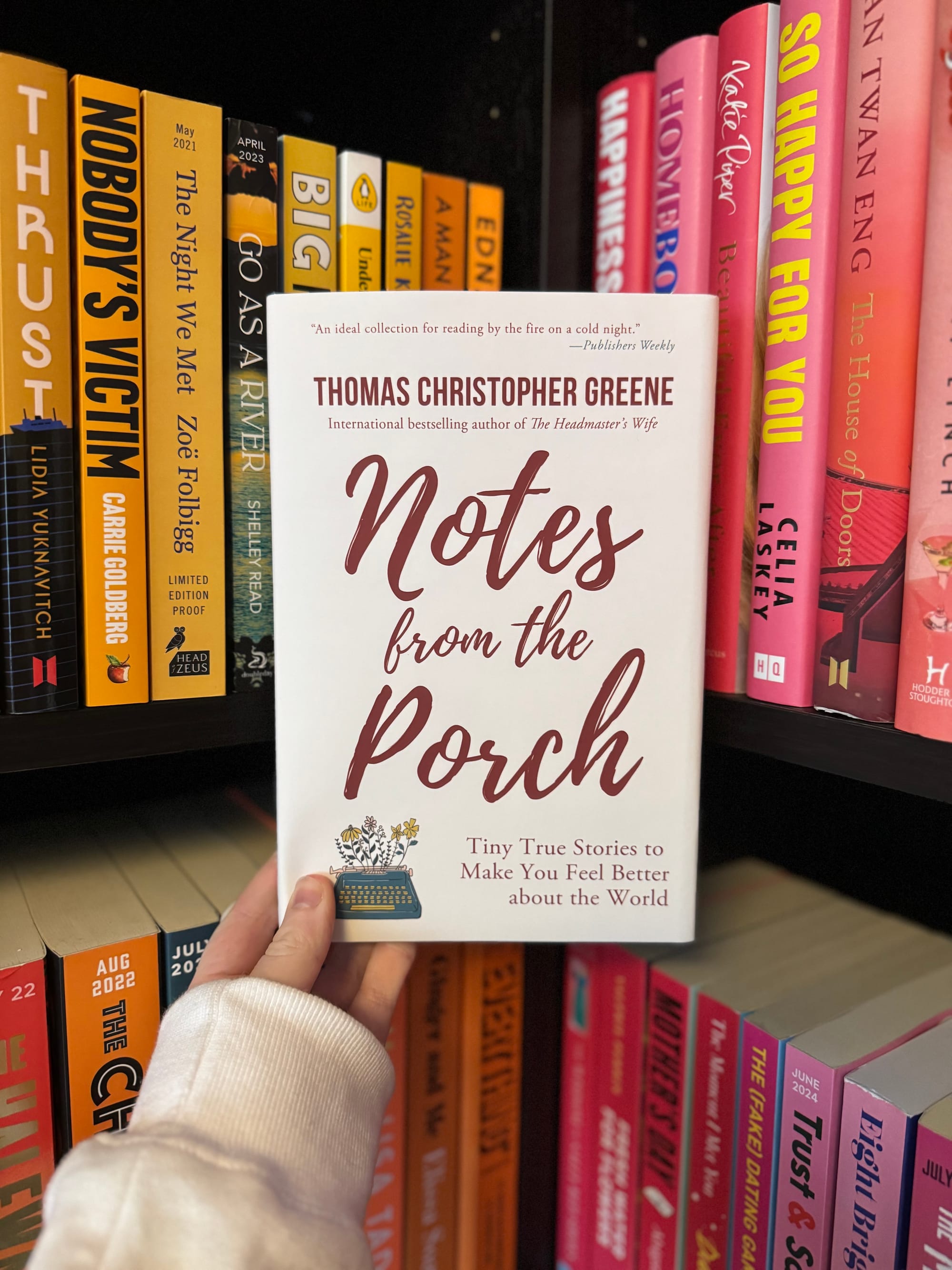 Person holding "Notes from the Porch" by Thomas Christopher Greene in a colorful bookstore. Bright book spines in yellow, pink, and orange.