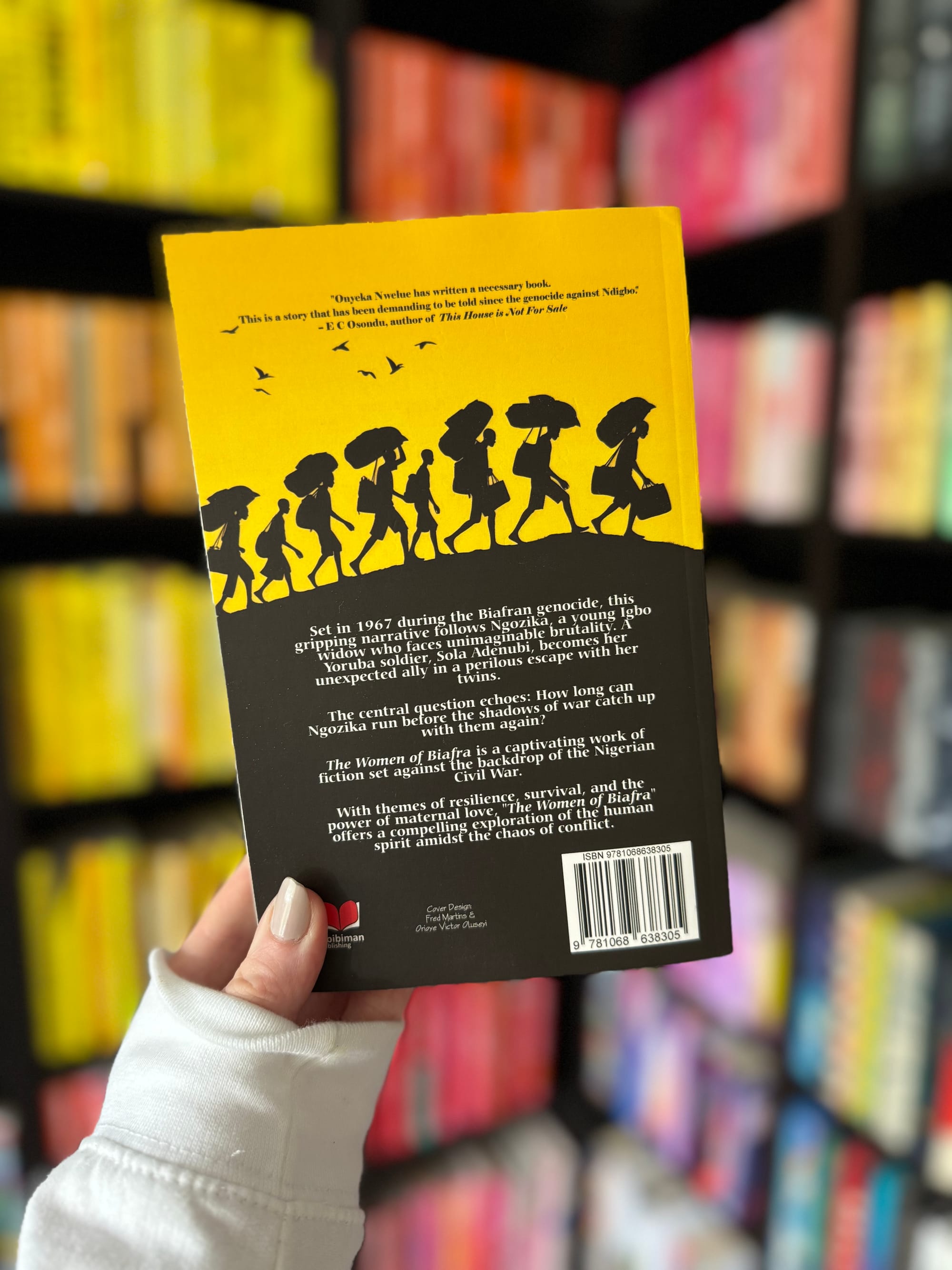 Person holding a book with a yellow and black cover, featuring silhouettes of people walking. Background of colorful, blurred bookshelves.