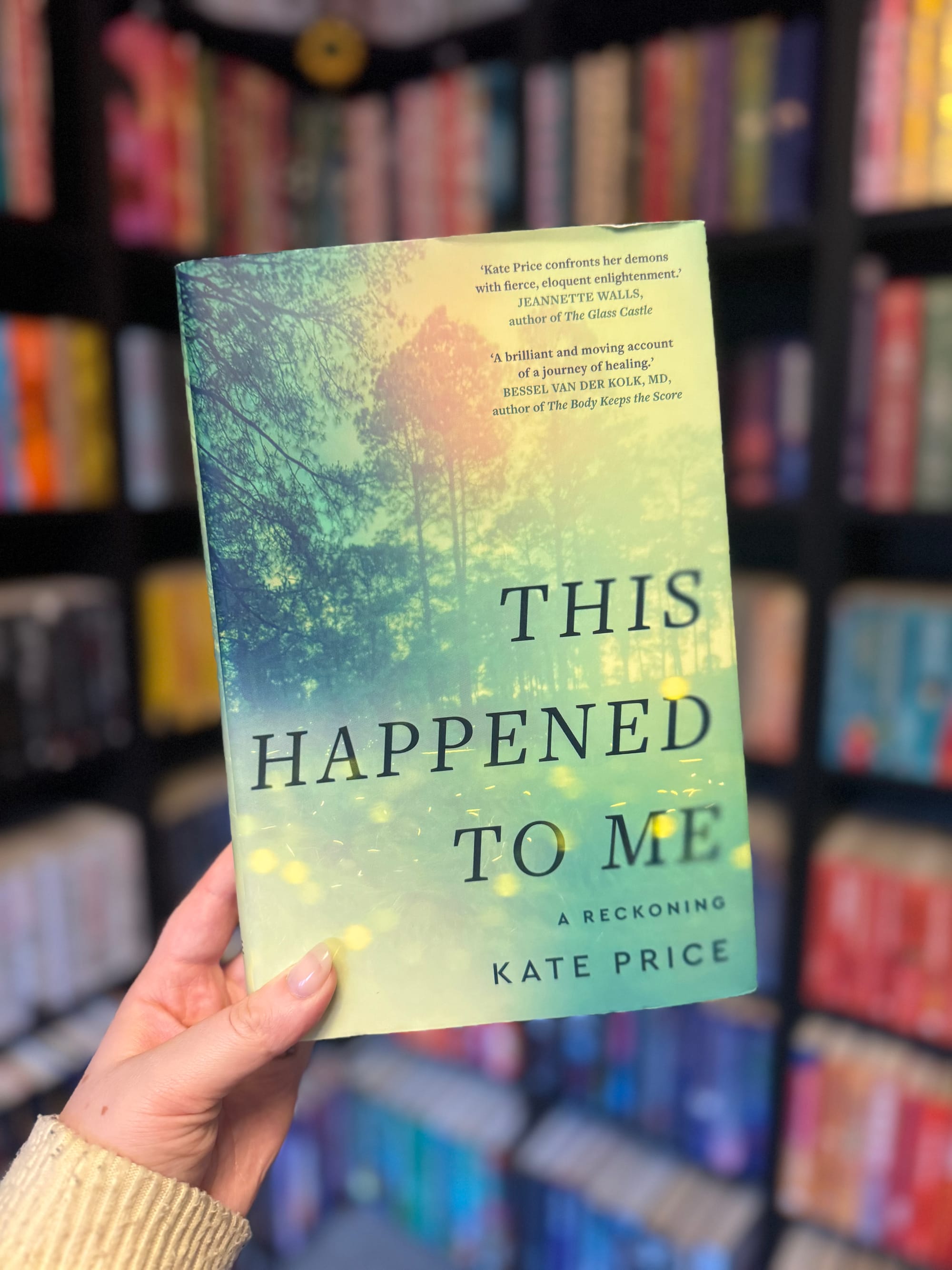 A hand holds "This Happened to Me" by Kate Price. The book has a green cover with a forest scene. Bookshelves blur in the background.