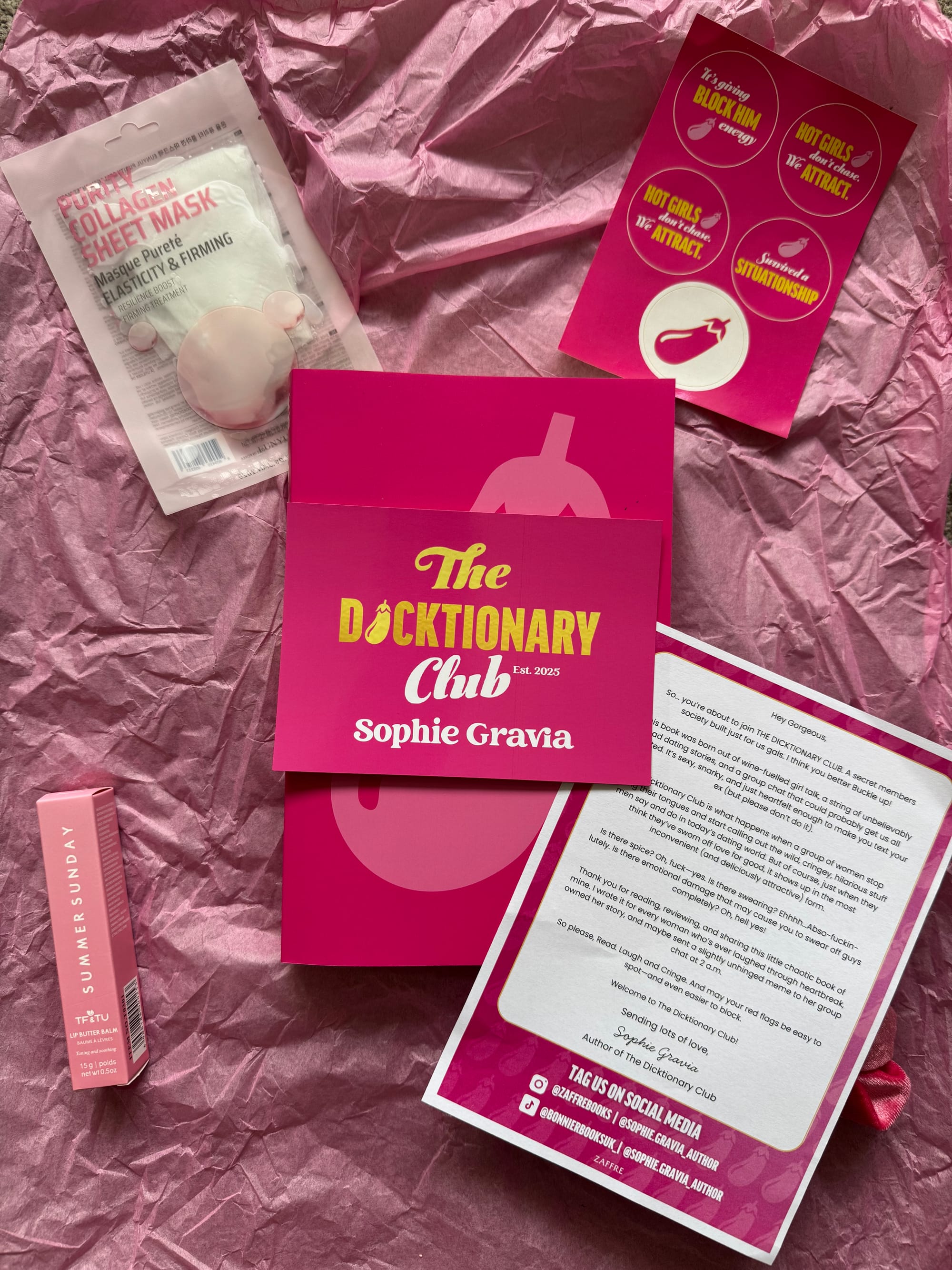 An open pink gift box with crinkled tissue paper lining the interior. Inside rests a rose-pink velvet scrunchie and a glossy card featuring gold text that reads: โThe Dicktionary Club Est. 2025 Sophie Gravia.โ The card is centrally placed, adding a touch of elegance. In the background, partially visible books with vibrant yellow and red spines sit neatly on a bookshelf, adding contrast and context.
