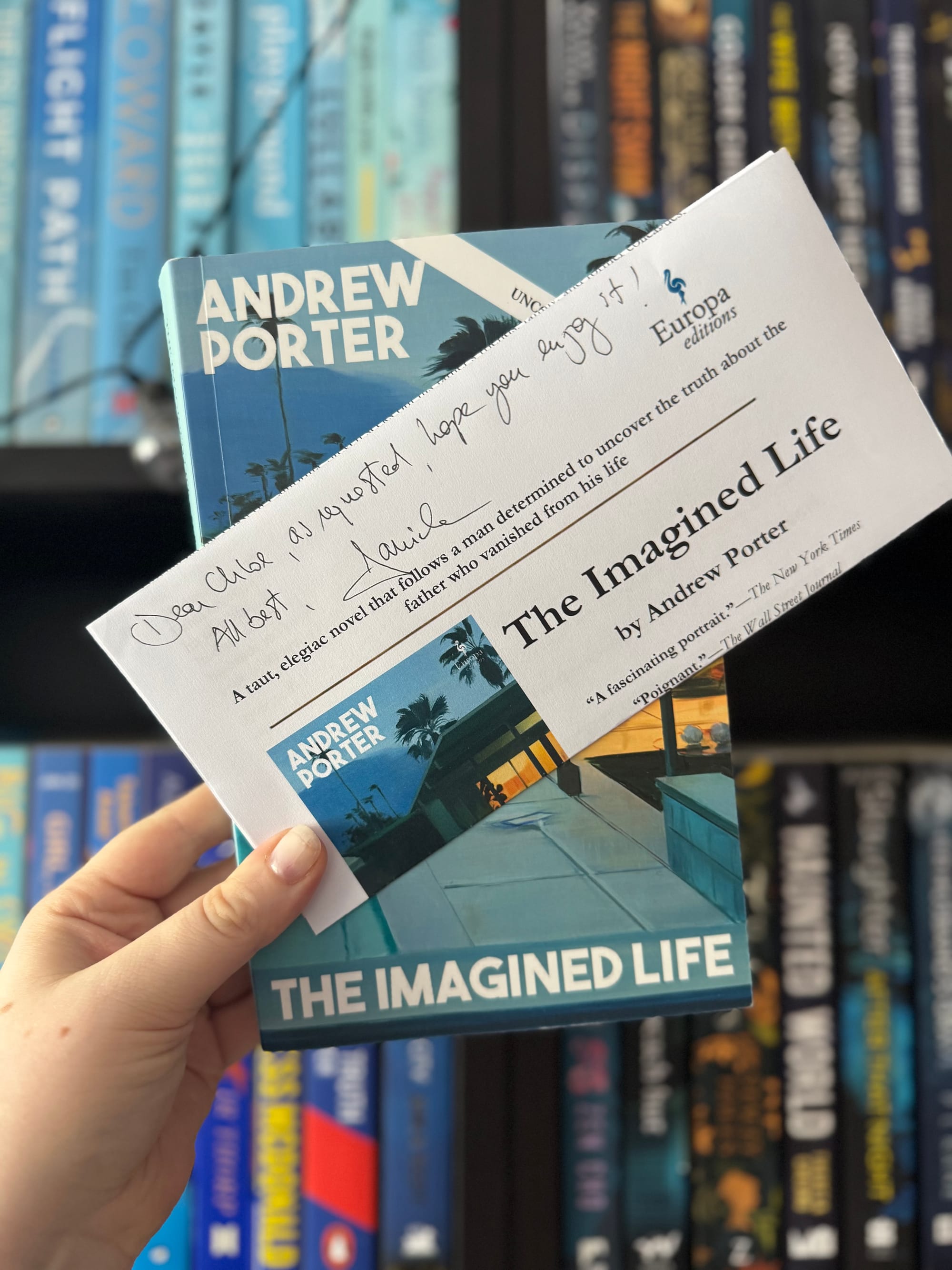 Hand holding "The Imagined Life" book by Andrew Porter with a handwritten note. Background: a bookshelf with colorful spines.