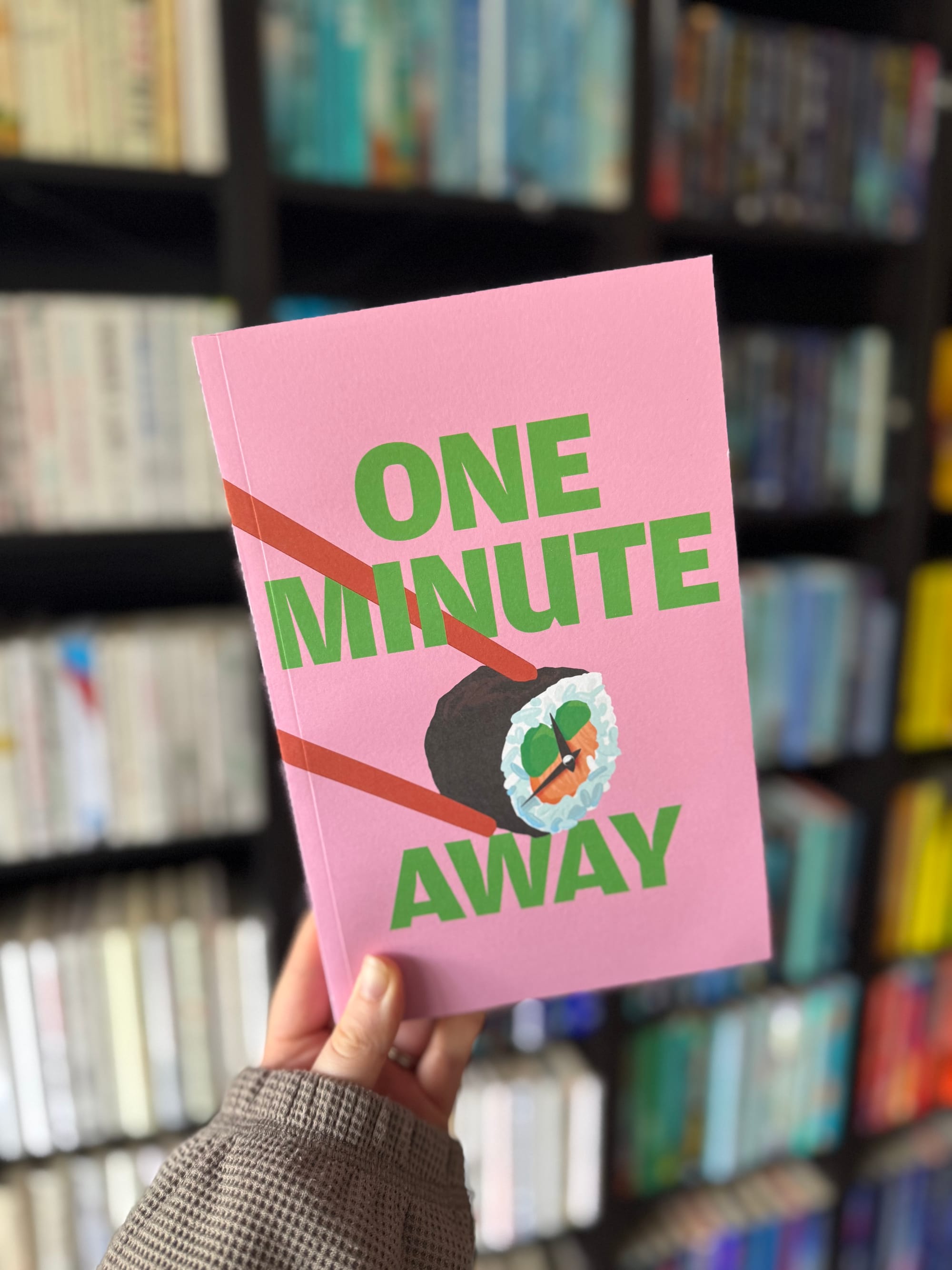 Hand holds a pink book titled "One Minute Away" with a sushi illustration, against a blurred bookshelf background.