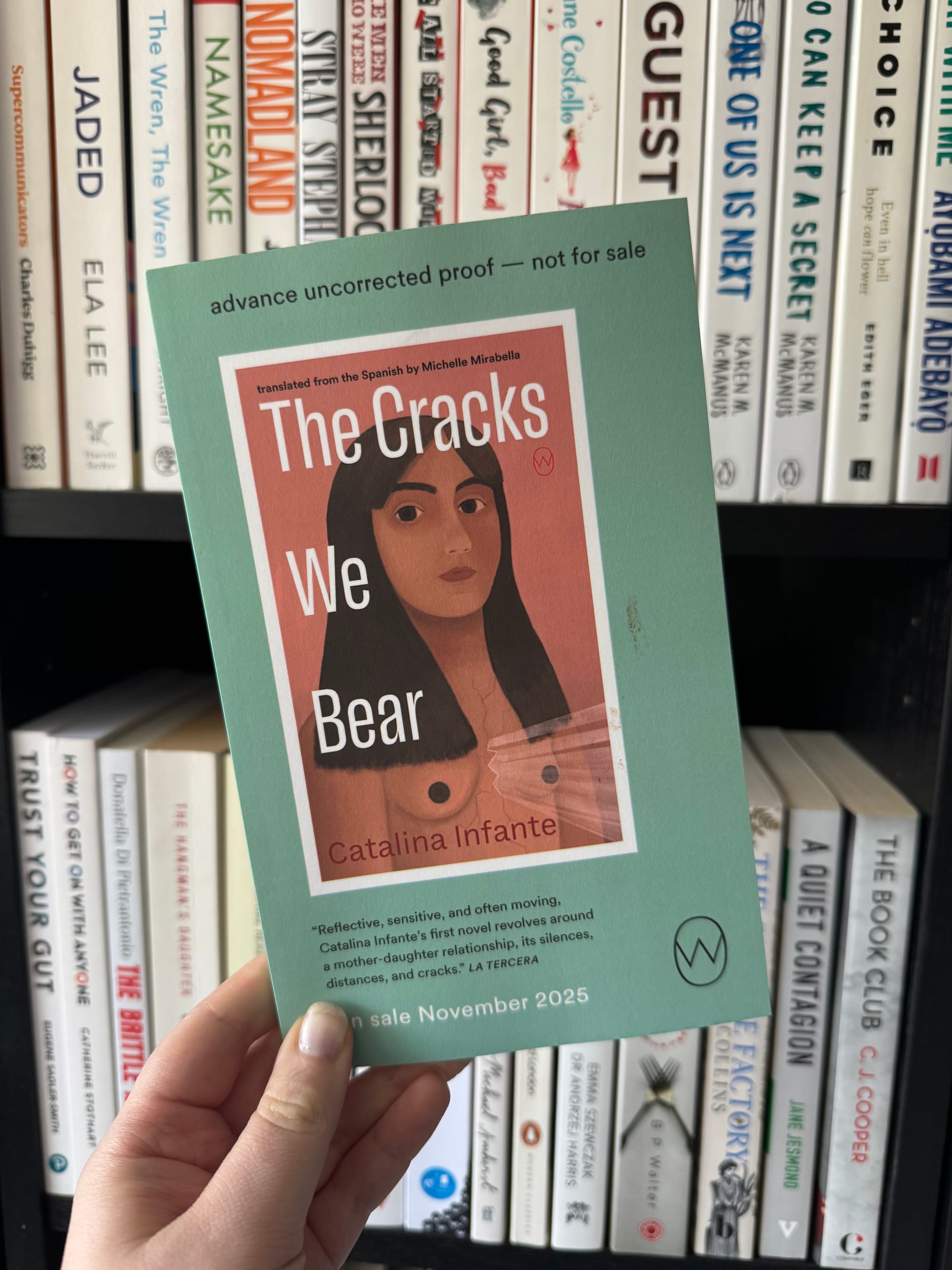 A hand holds a book titled "The Cracks We Bear" by Catalina Infante. Green cover with a woman's face. Bookshelf in background.