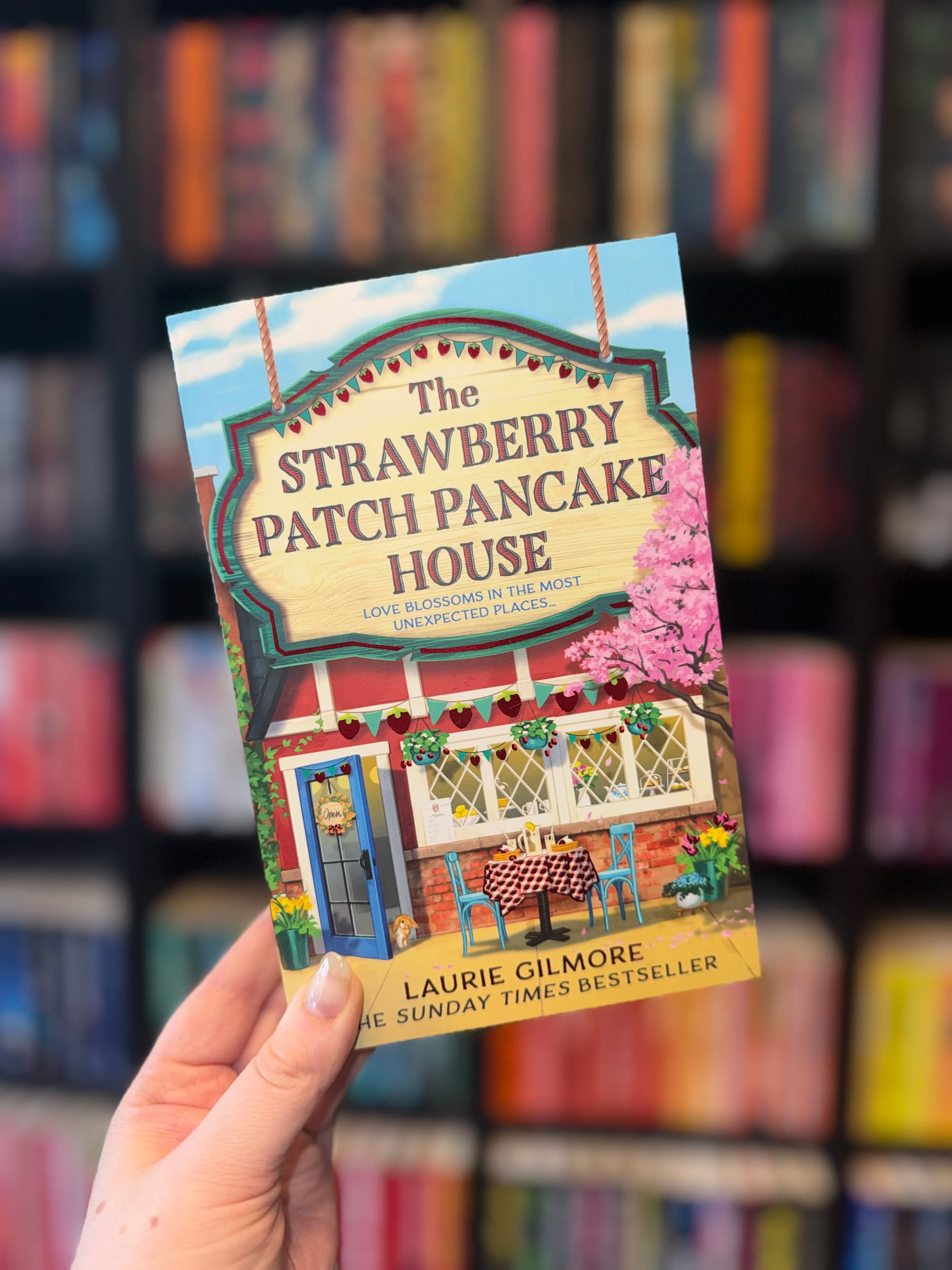 Hand holding "The Strawberry Patch Pancake House" book with colorful cover featuring a cozy café scene. Blurred bookshelves in background.
