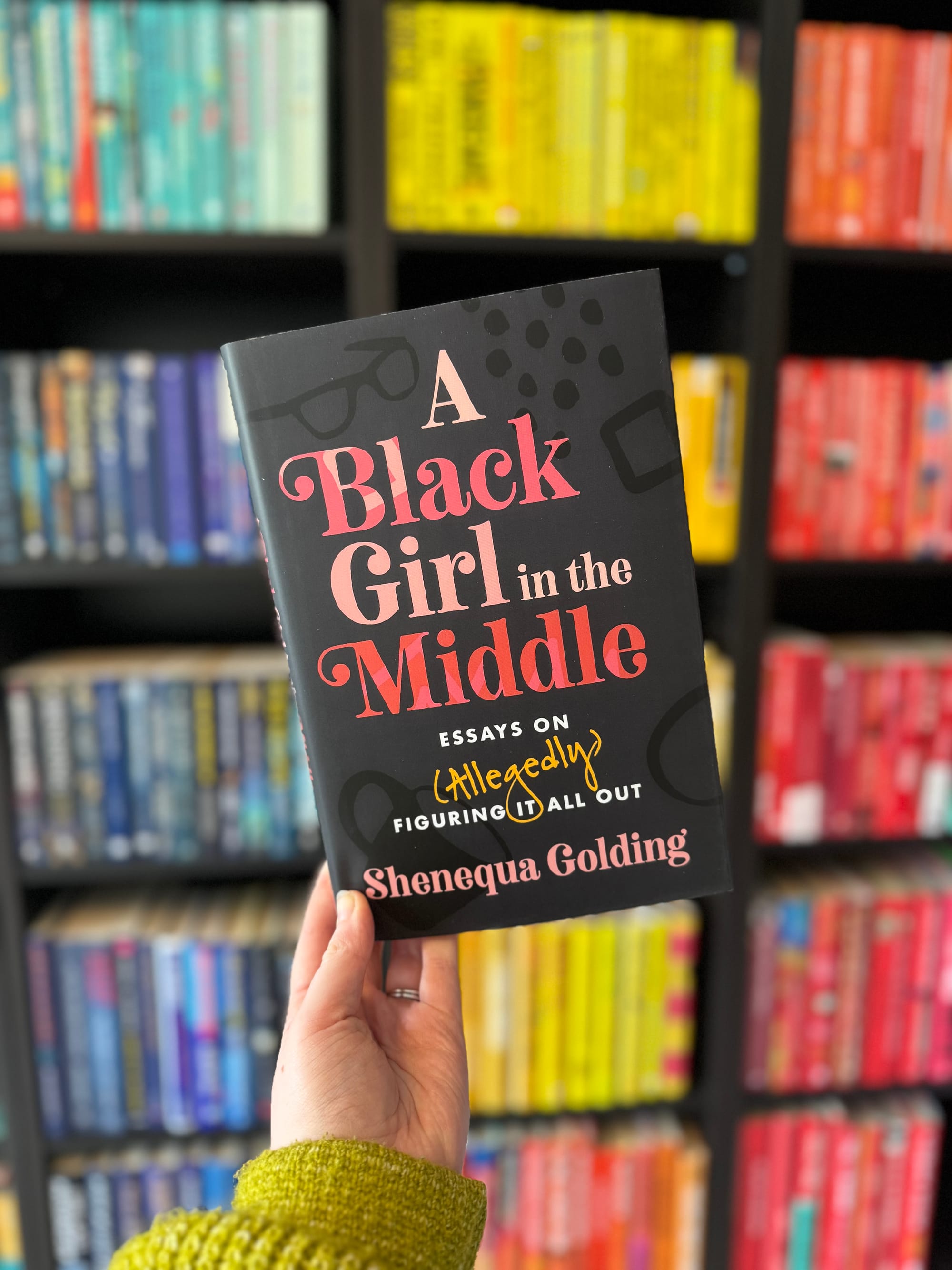 Hand holds "A Black Girl in the Middle" book against rainbow-colored bookshelf. The book's title is prominent, creating an intriguing focal point.