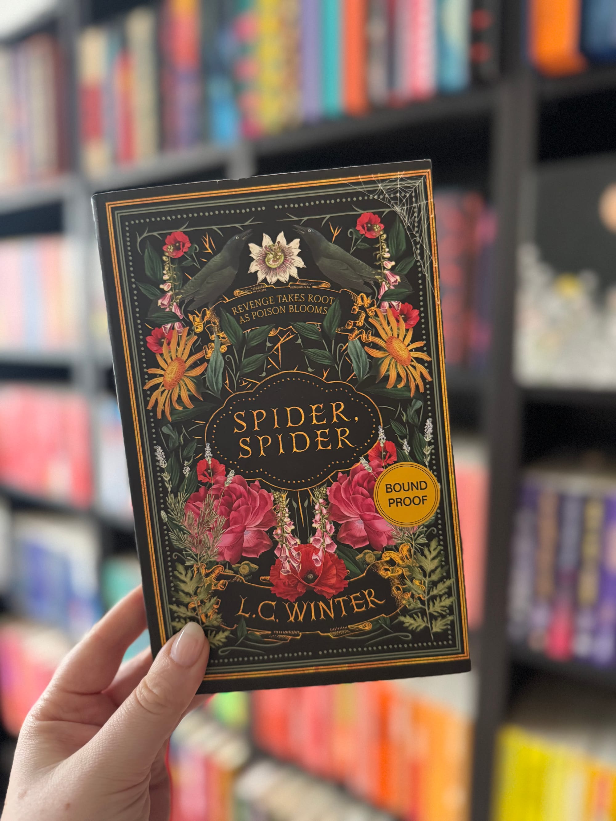 A hand holding the book "Spider, Spider" by L.C. Winter. Dark floral cover with crows and text "Revenge takes root as poison blooms." Bookshelf backdrop.