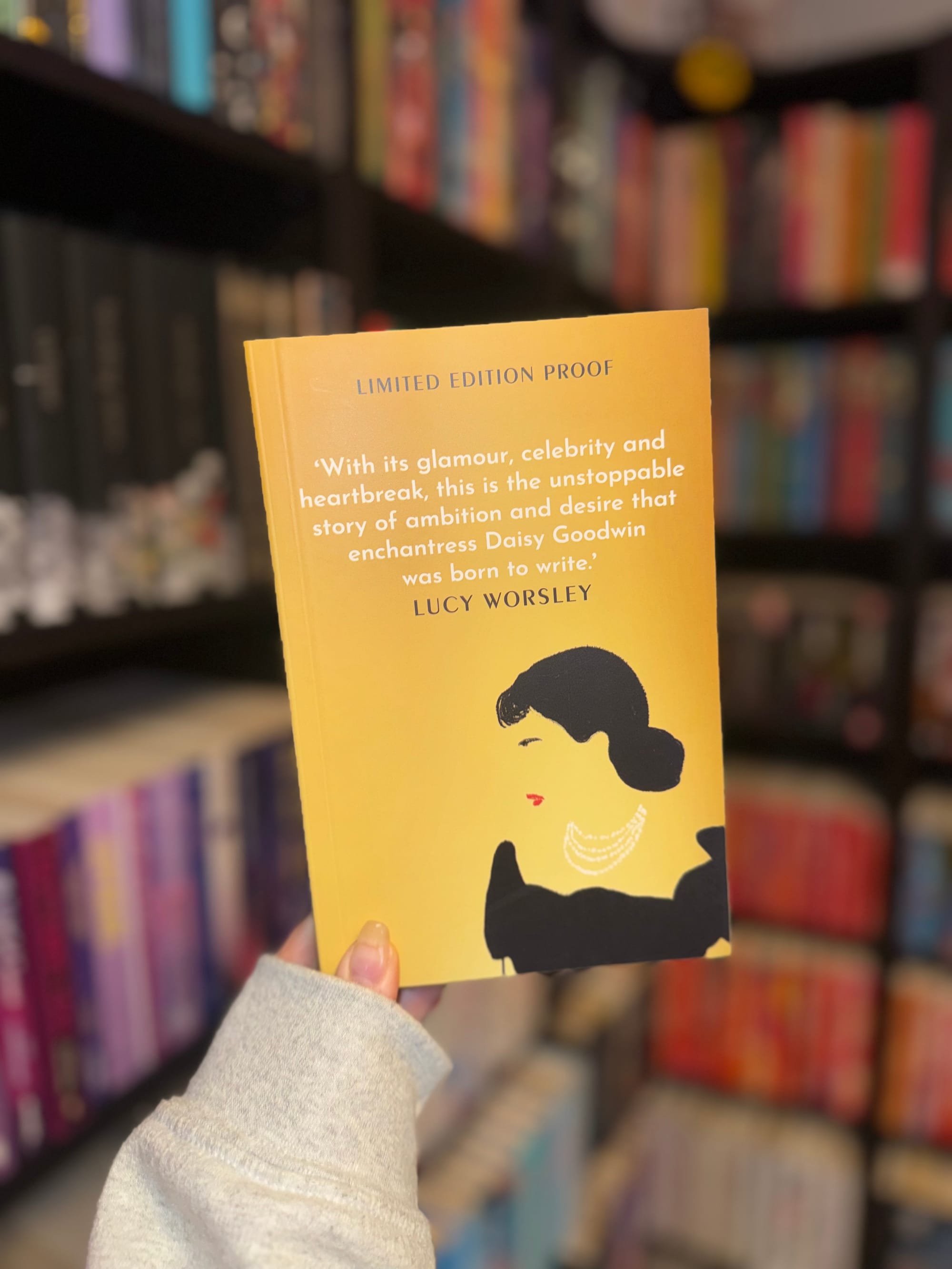 A hand holds a yellow book titled "Limited Edition Proof" with a woman's silhouette. Bookshelves with colorful spines are blurred in the background.