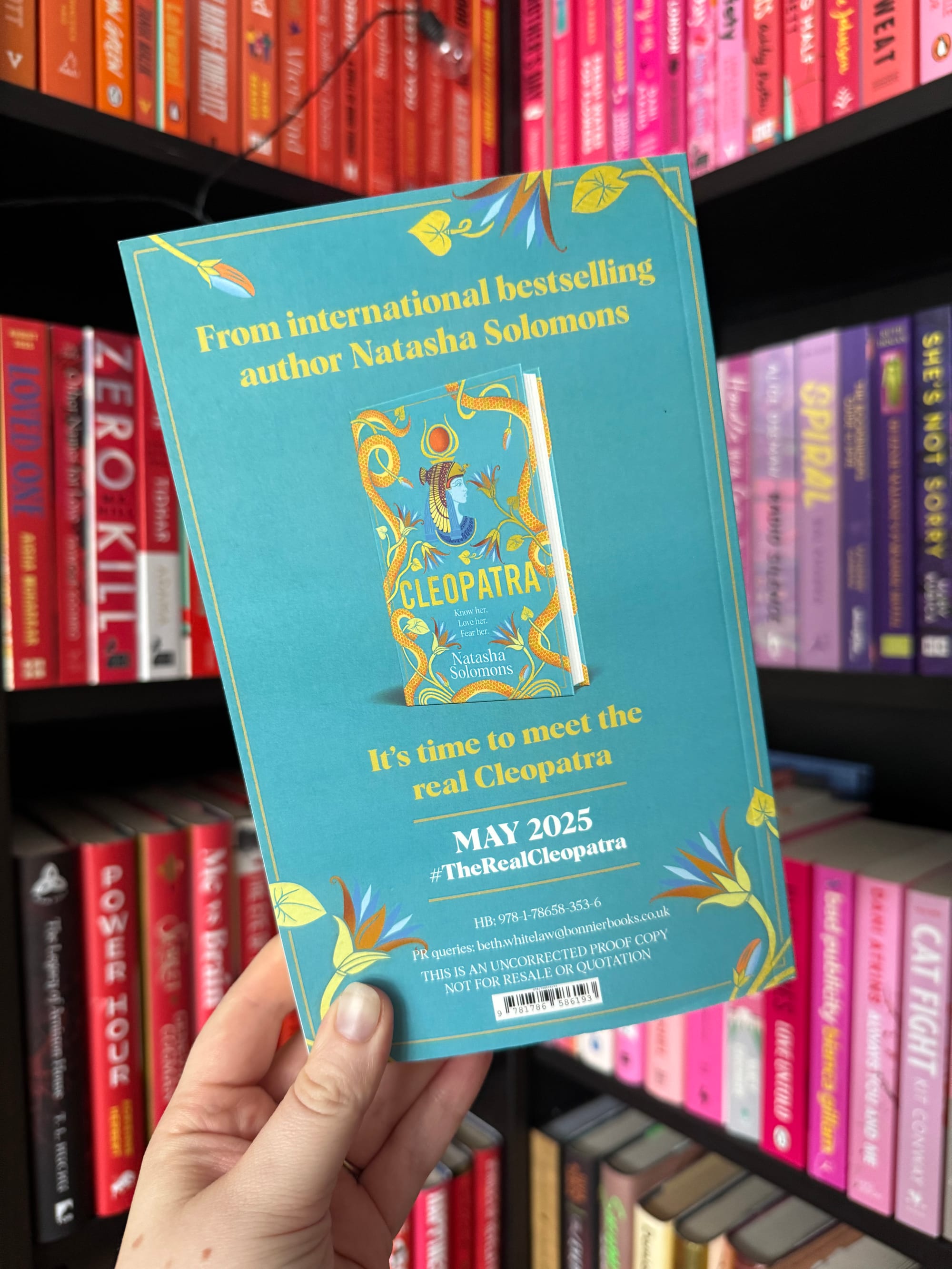 A hand holds a blue book flyer for "Cleopatra" by Natasha Solomons. Text reads "May 2025" against a colorful bookshelf backdrop.