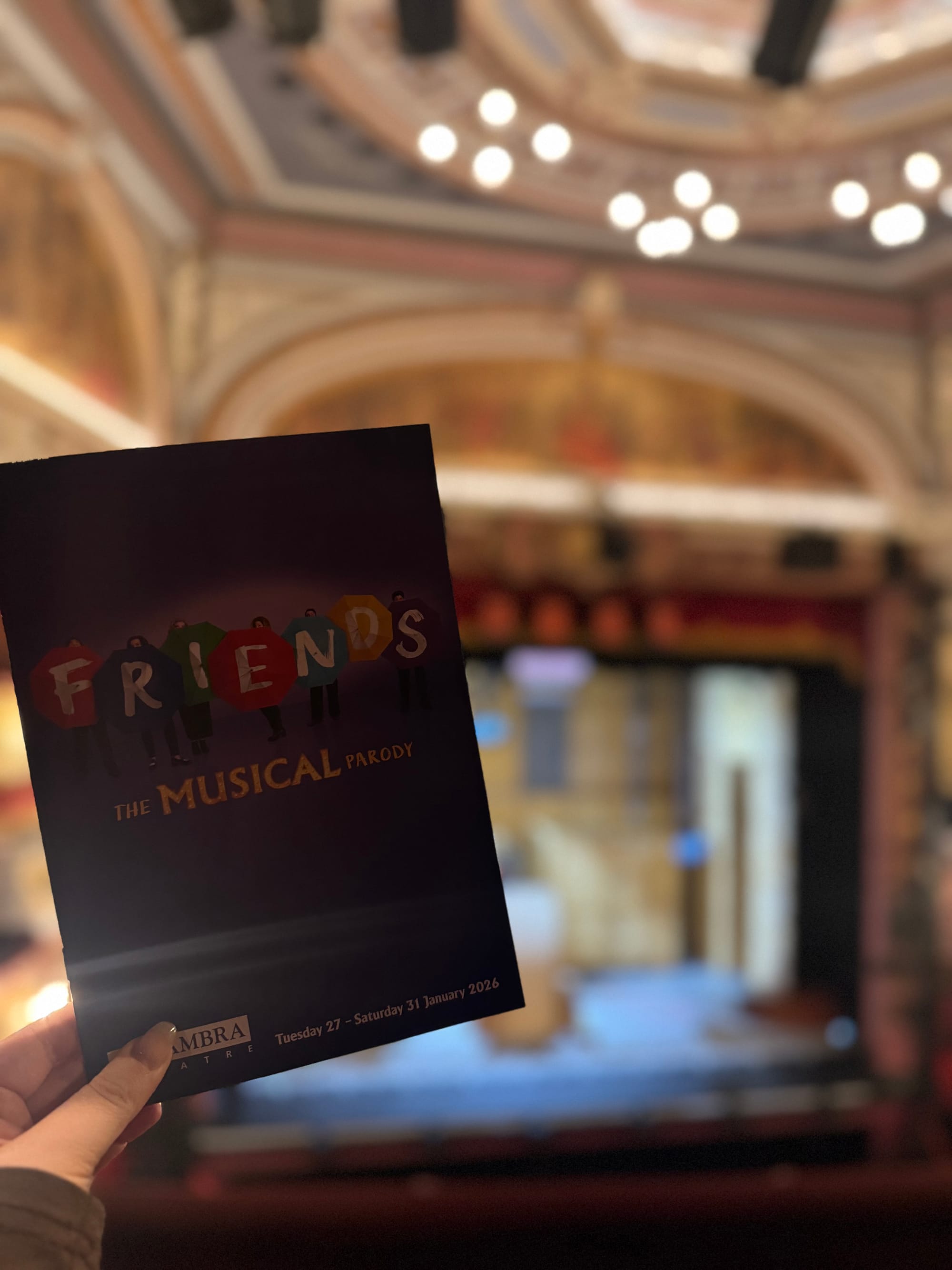 Programme for FRIENDS! The Musical Parody held up inside the Alhambra Theatre in Bradford, with the stage and ornate auditorium softly blurred in the background.