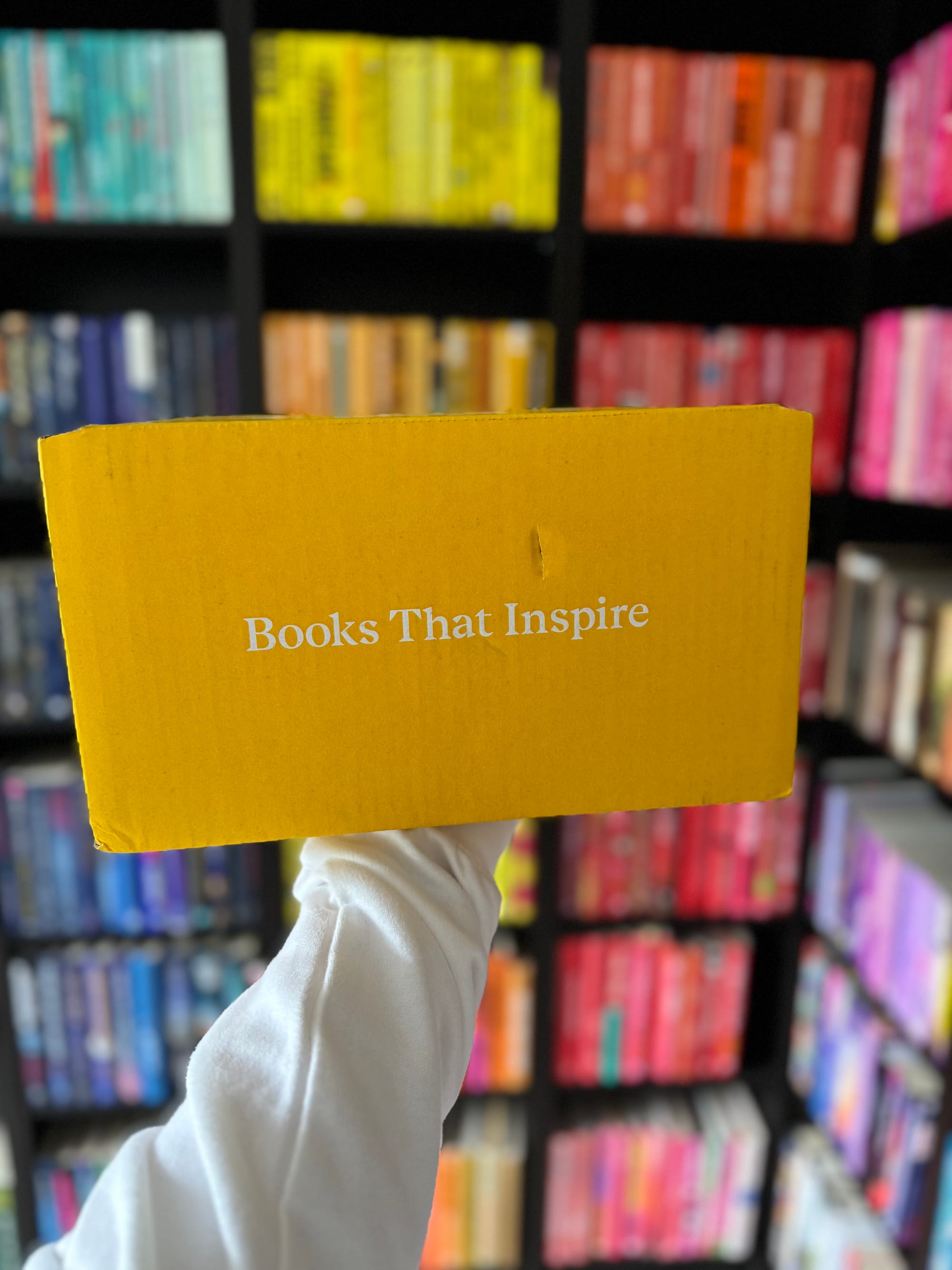 A hand holds a yellow box with "Books That Inspire" text, against a colorful bookshelf background. The mood is uplifting and vibrant.