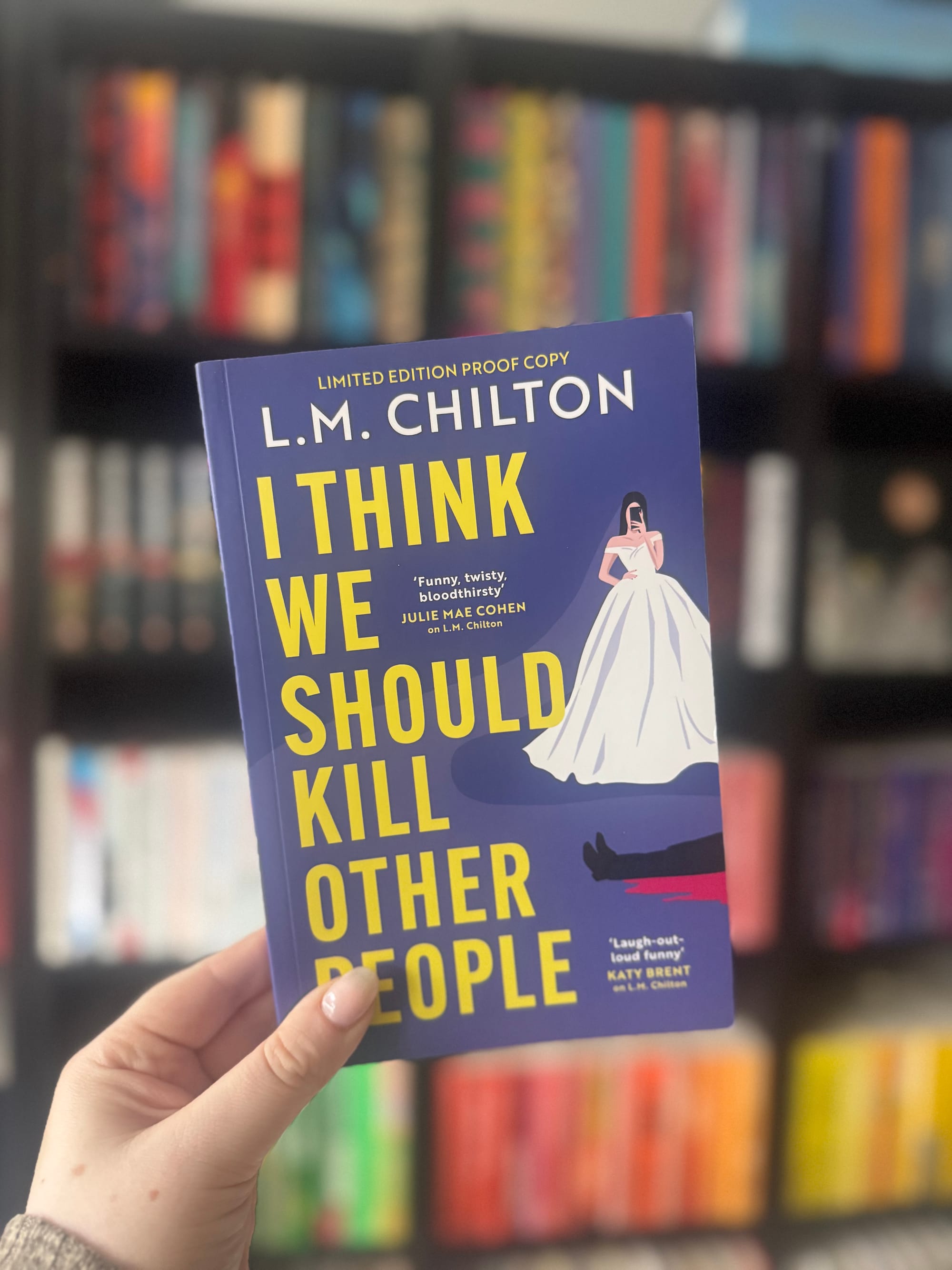 Hand holding a book titled "I Think We Should Kill Other People" by L.M. Chilton. Background shows colorful, blurred books on a shelf.