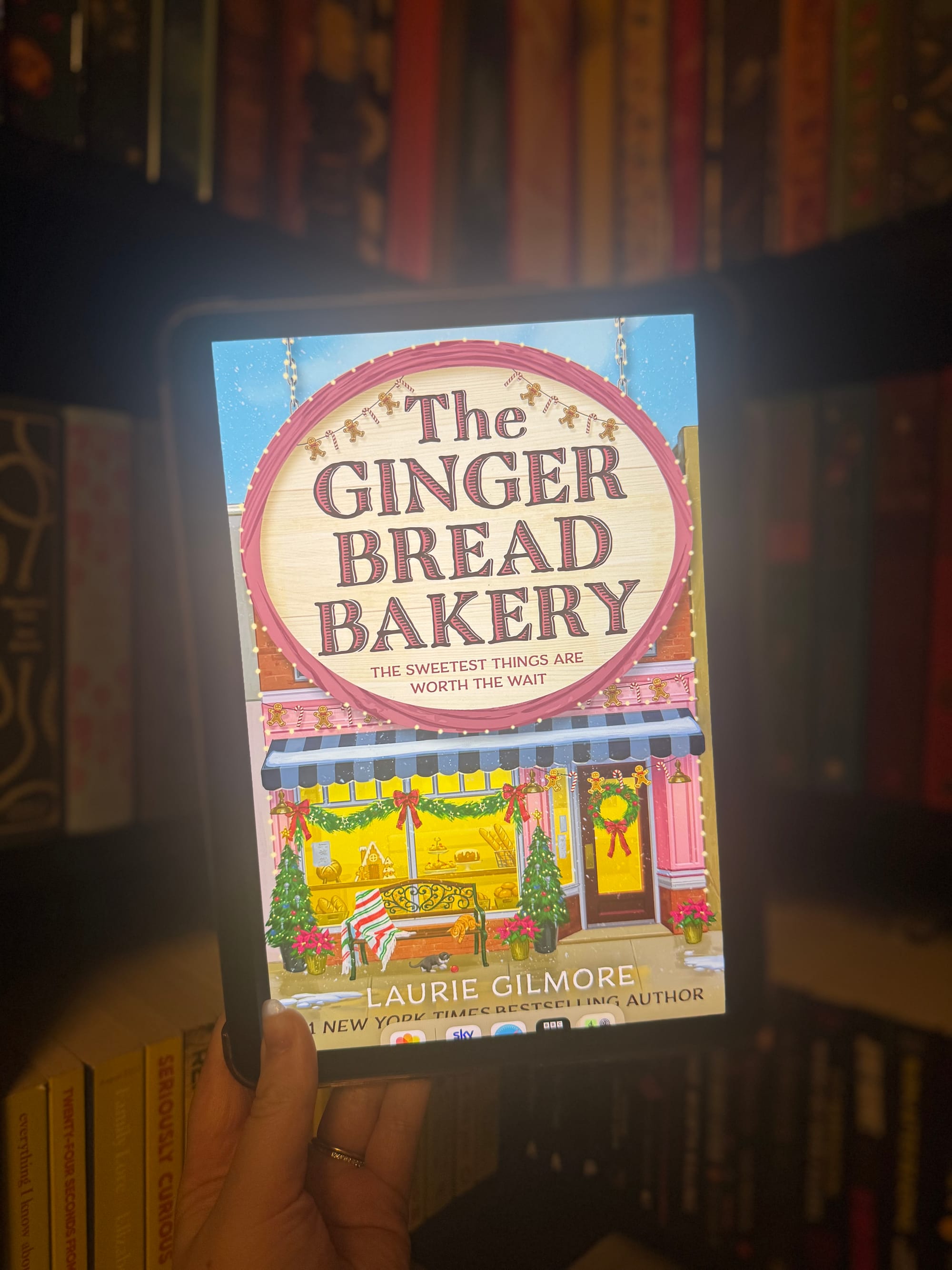 Hand holding a tablet displaying "The Gingerbread Bakery" cover with festive bakery illustration. Background shows blurred books on shelves.
