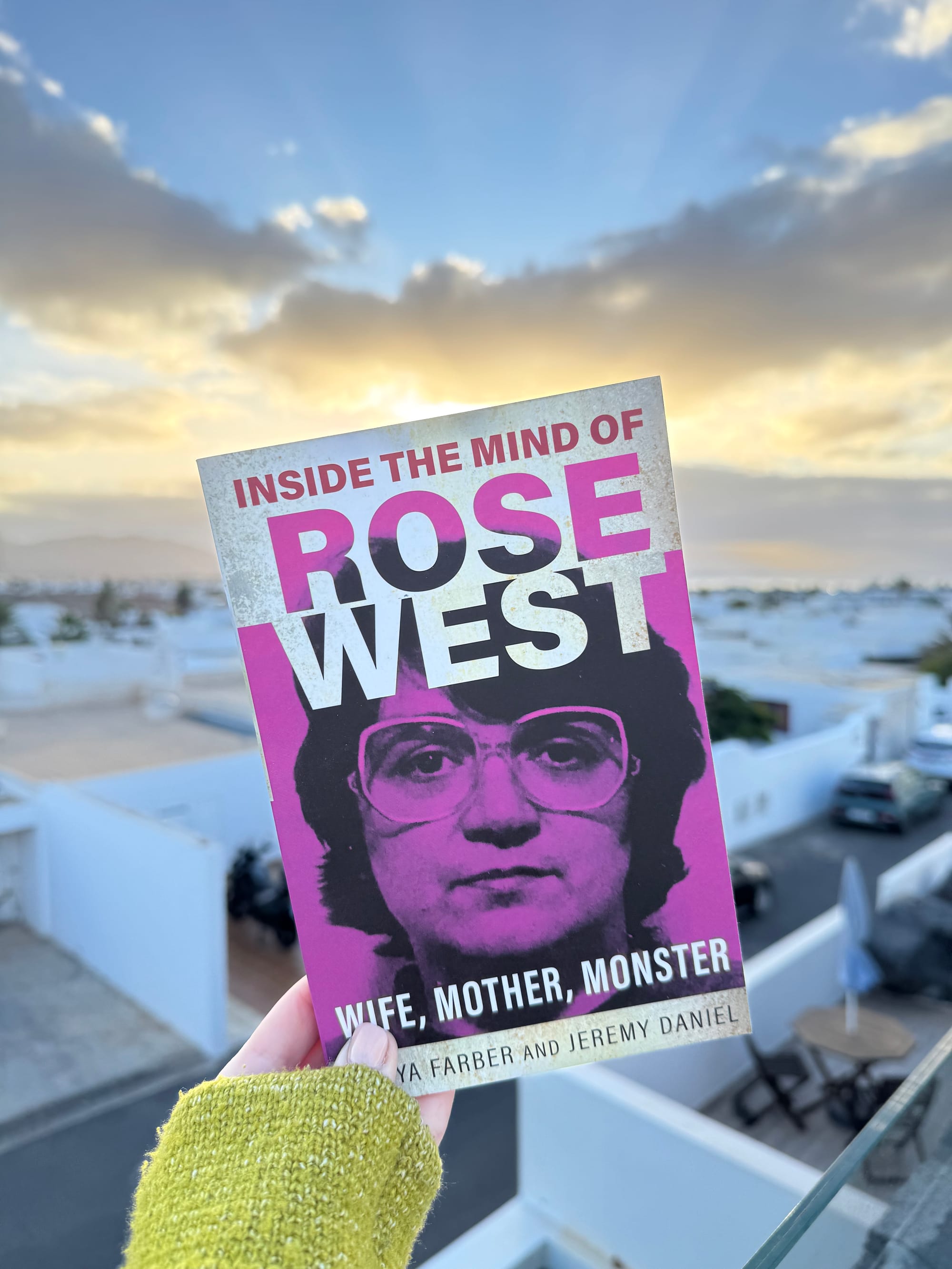 Person holds the book "Inside the Mind of Rose West" against a sunset backdrop. Purple cover with bold text and a serious portrait of Rose West. 