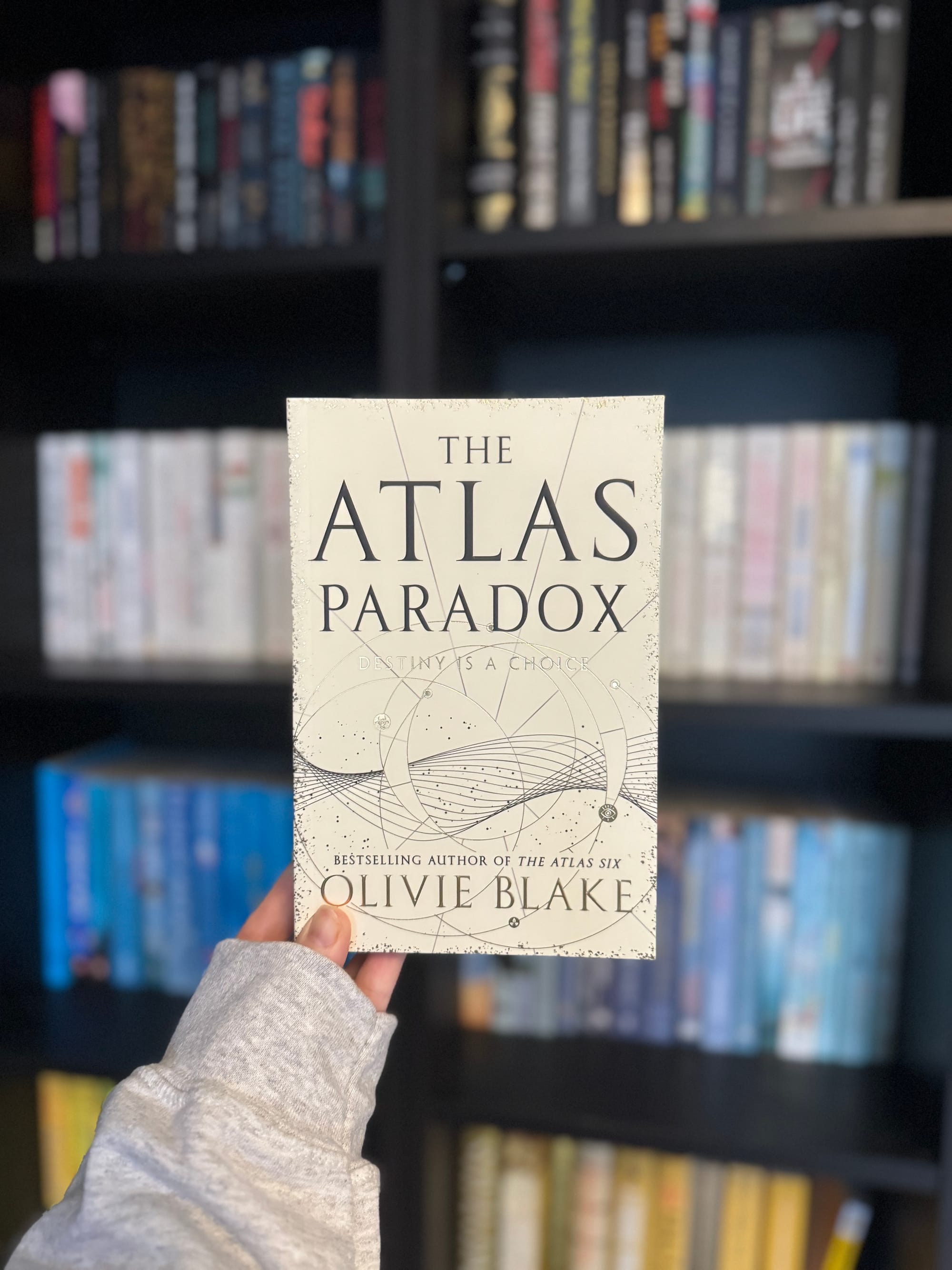 A hand holds the book "The Atlas Paradox" by Olivie Blake against a blurred bookshelf. The cover is cream with cosmic designs.