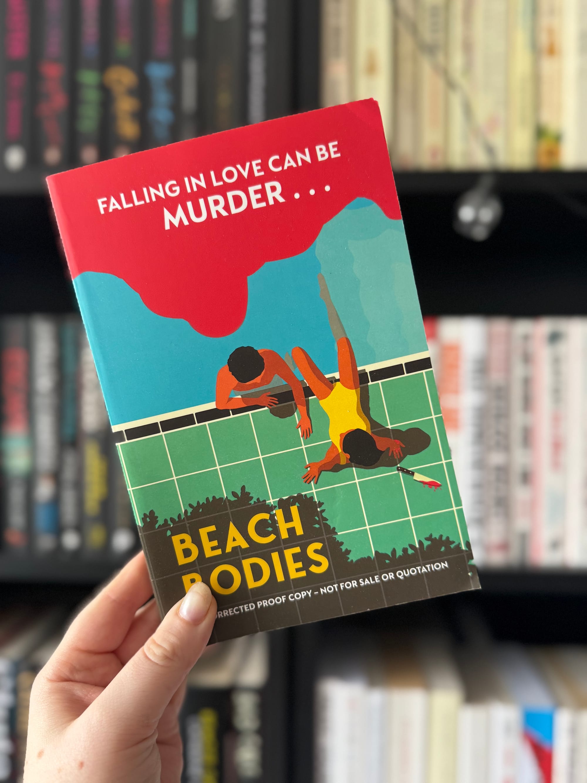 Hand holding a book titled "Beach Bodies." Cover shows two people by a pool. Bold text reads "FALLING IN LOVE CAN BE MURDER..." Background: bookshelf.