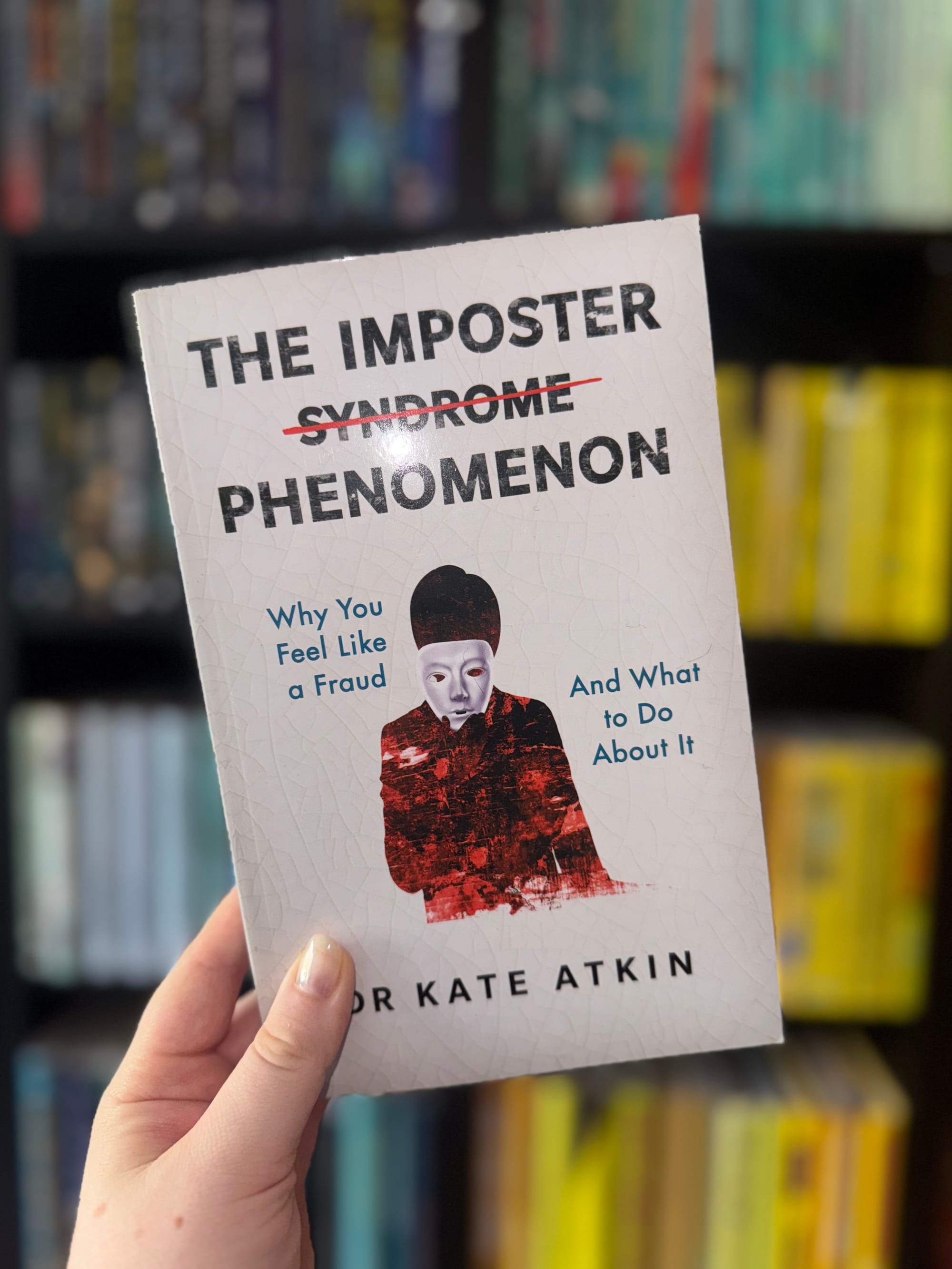 Close-up of a hand holding the book "The Imposter Phenomenon" by Dr. Kate Atkin, in front of a blurred bookshelf. The cover features a masked figure.