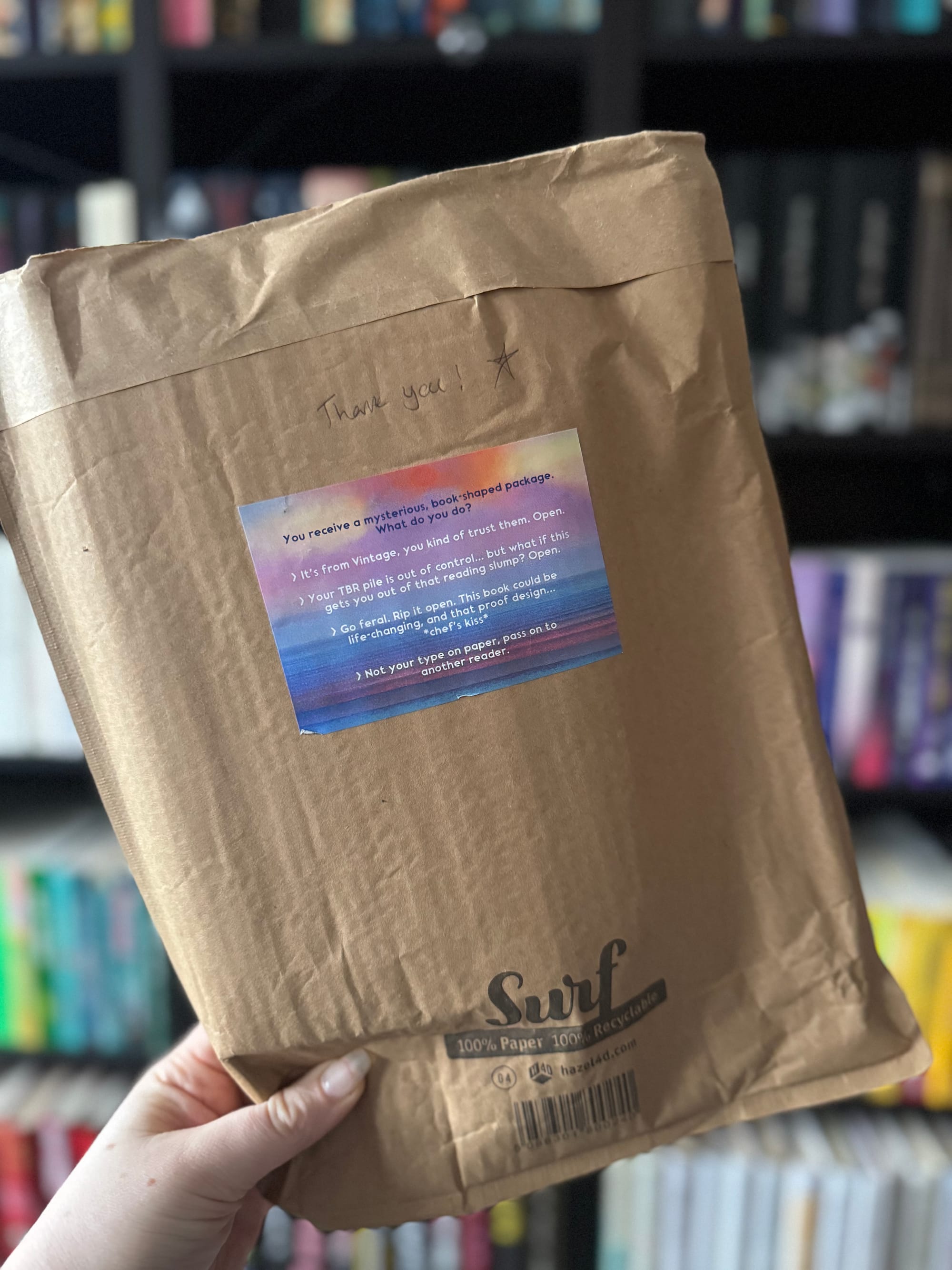 A hand holding a brown paper parcel with a handwritten thank you and a star on the front. A colourful sticker with printed text is attached to the parcel. Bookshelves filled with books are blurred in the background.