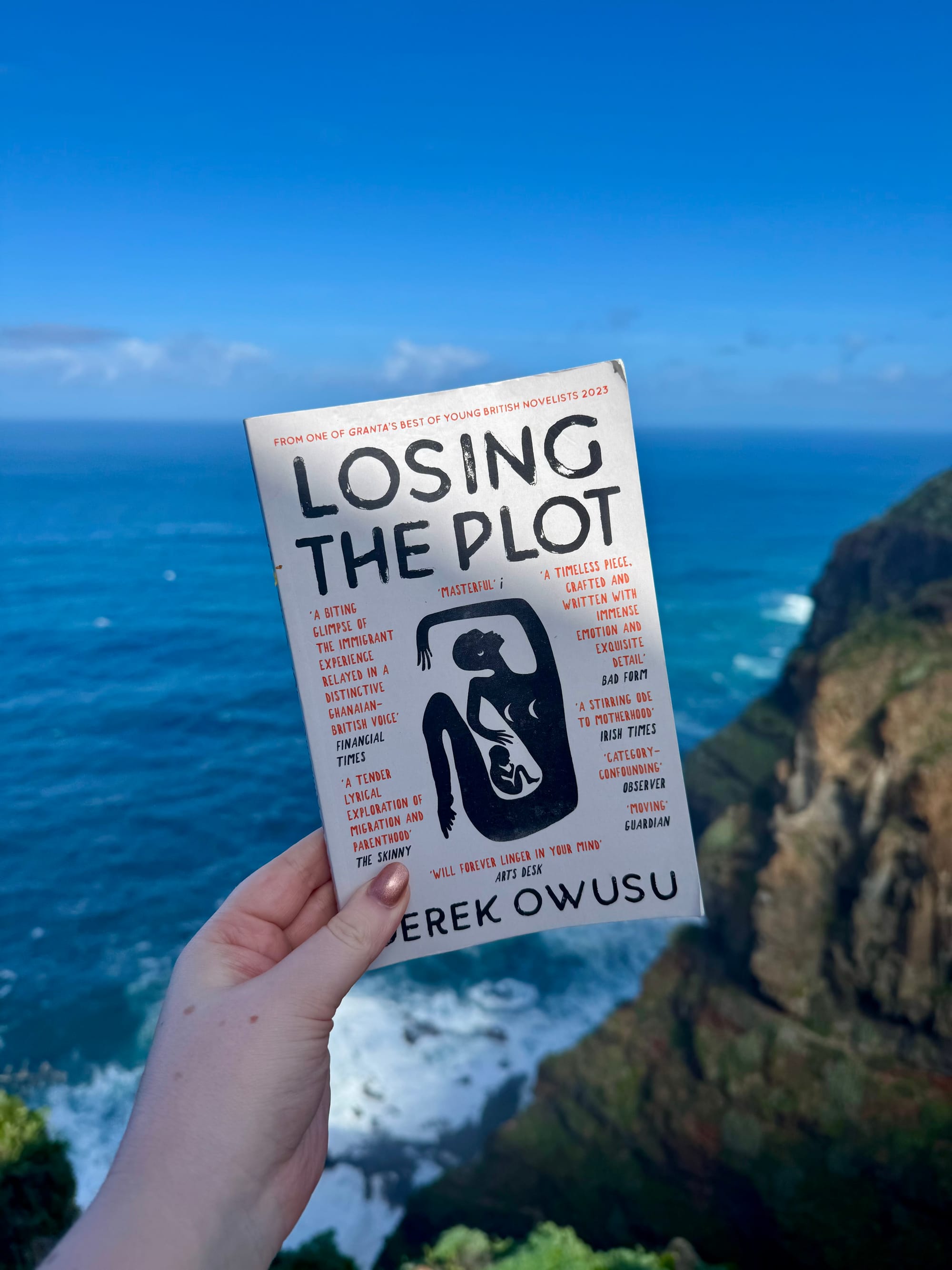 Hand holding the book "Losing the Plot" by Derek Owusu against a backdrop of ocean cliffs and blue sky.