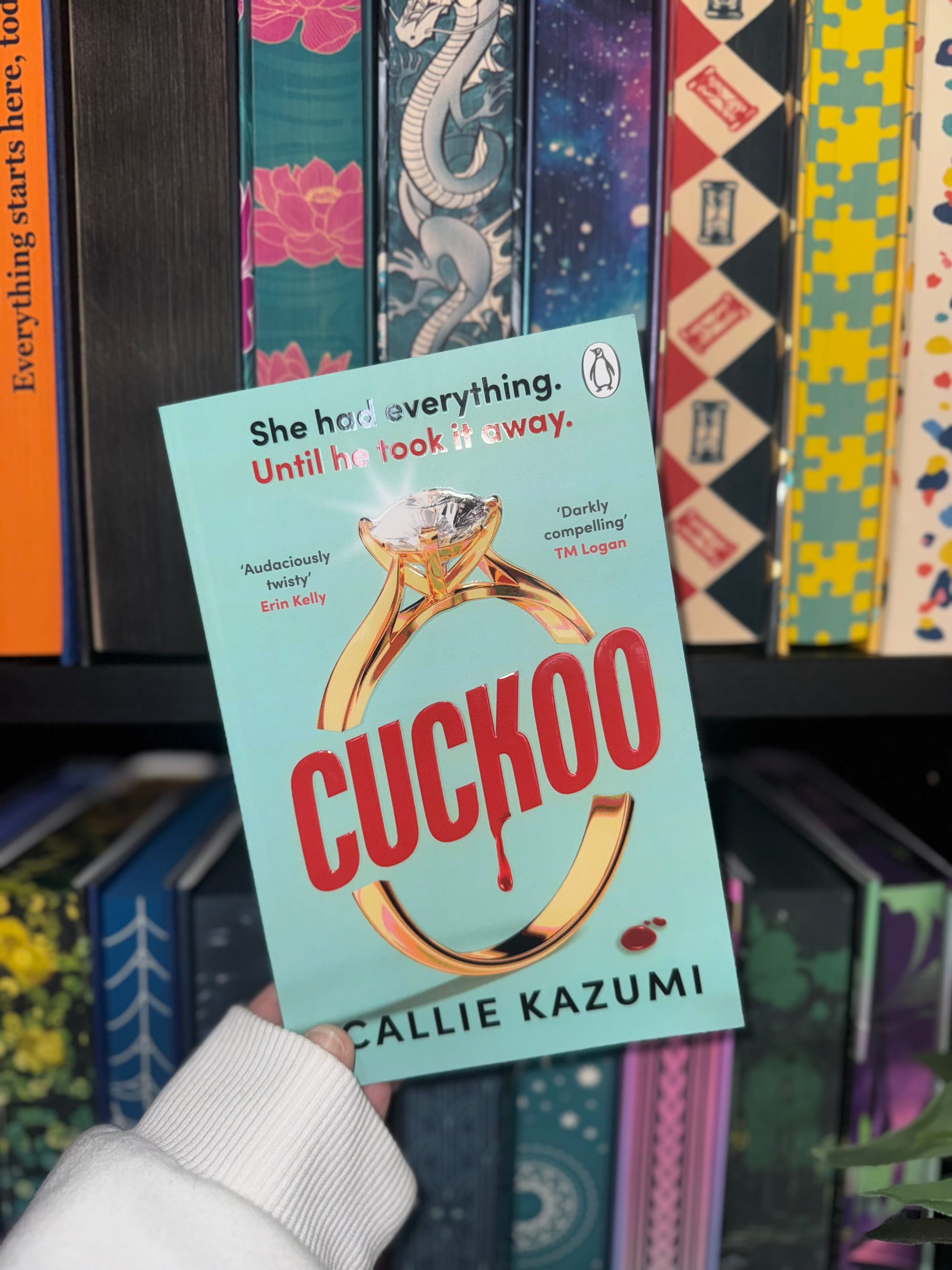Person holds "Cuckoo" book in front of colorful bookshelf. Cover text: "She had everything. Until he took it away." Bright and intriguing.