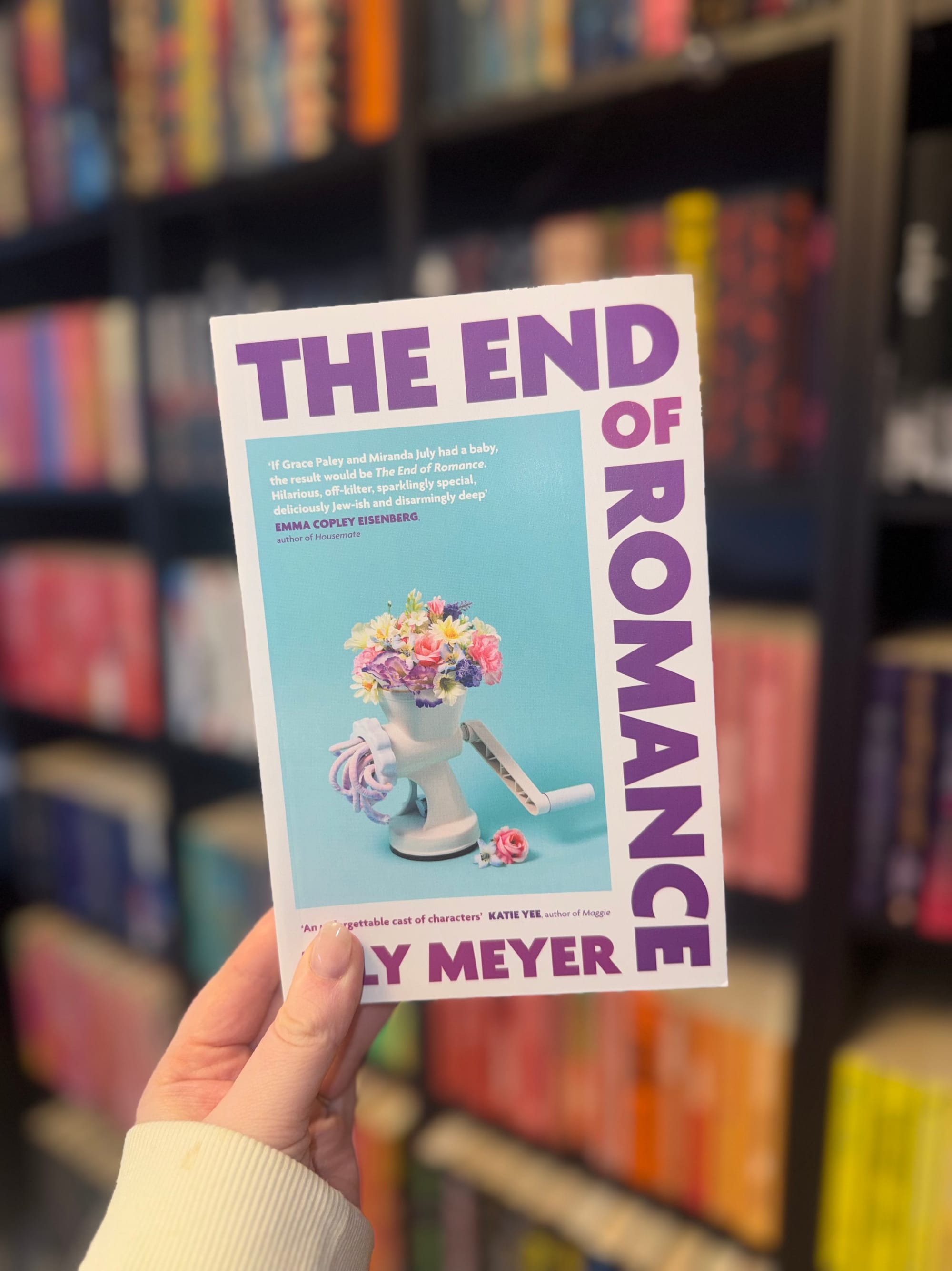 Hand holding a book titled "The End of Romance" by Lily Meyer, featuring a flower-filled grinder on a blue background. Blurred bookshelves behind.