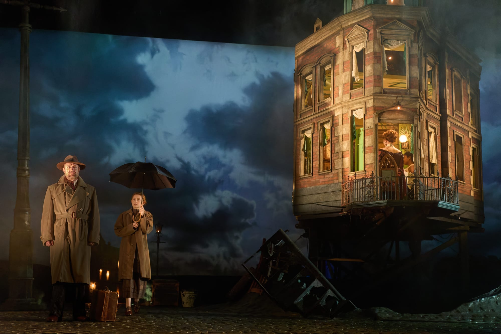 Two people in coats stand under an umbrella by a lamppost. A man and woman are visible in a house. Dark, cloudy sky creates a moody atmosphere.
