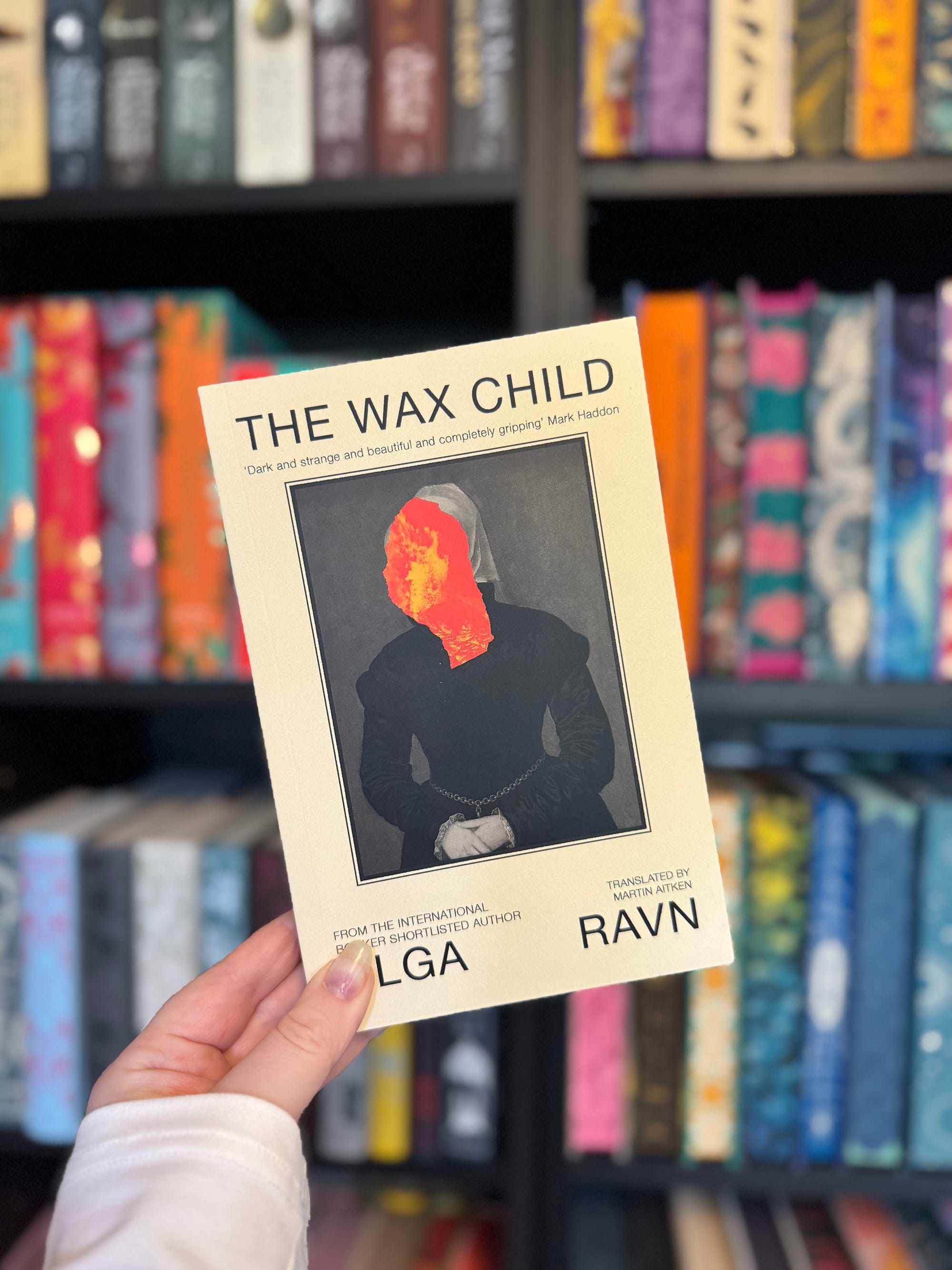 A hand holds "The Wax Child" book with a fiery face on the cover, set against colorful bookshelves in a library.