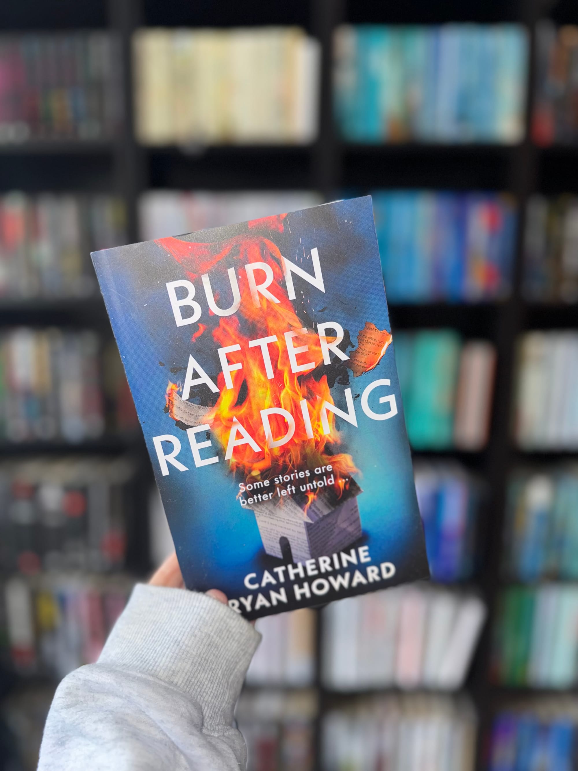 Hand holds a book titled "Burn After Reading" by Catherine Ryan Howard against a blurred bookshelf. Cover features flames and paper.