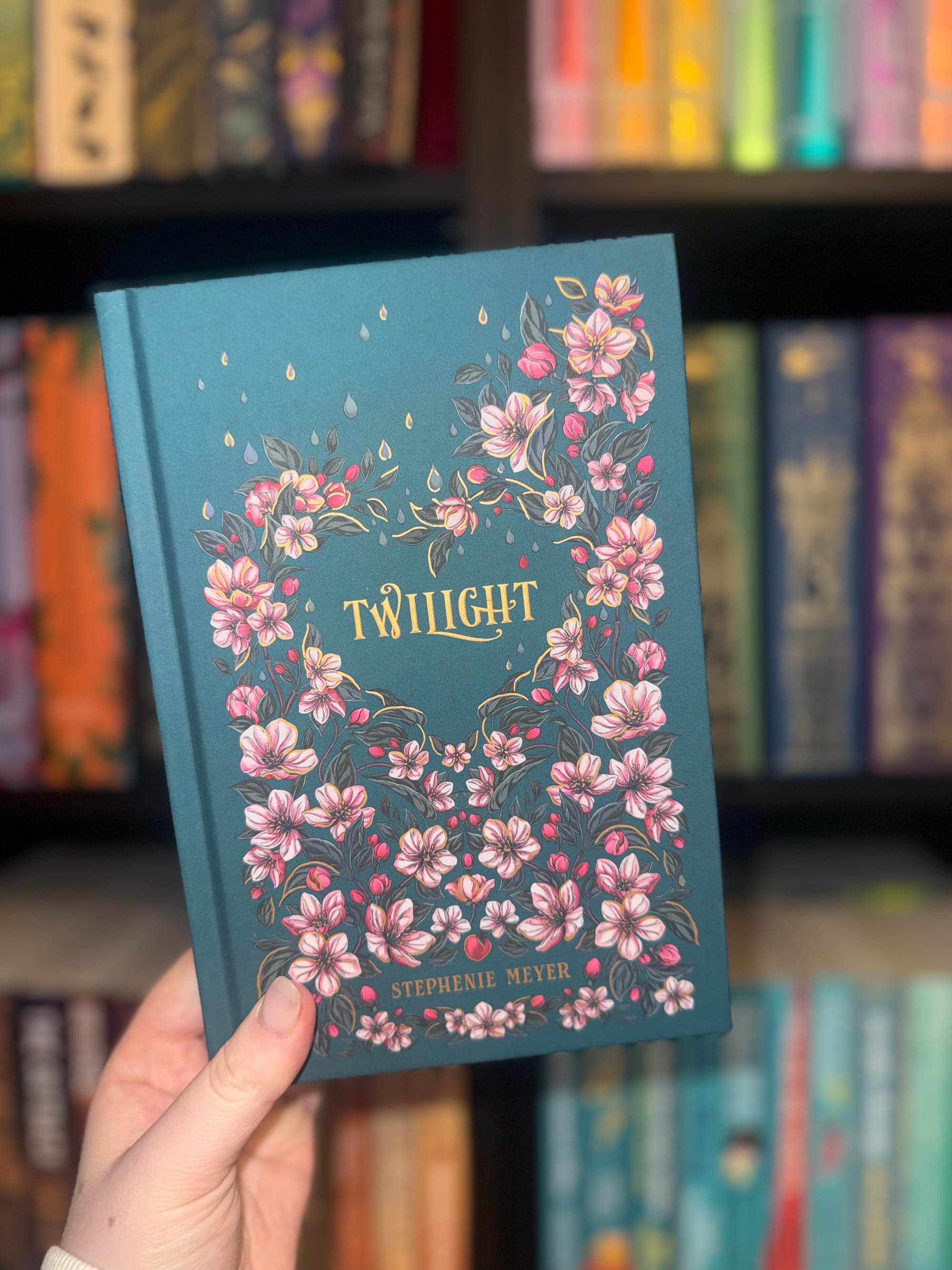 Hand holding a teal book with pink flowers, titled "Twilight" by Stephenie Meyer. Colorful books on a shelf are blurred in the background.