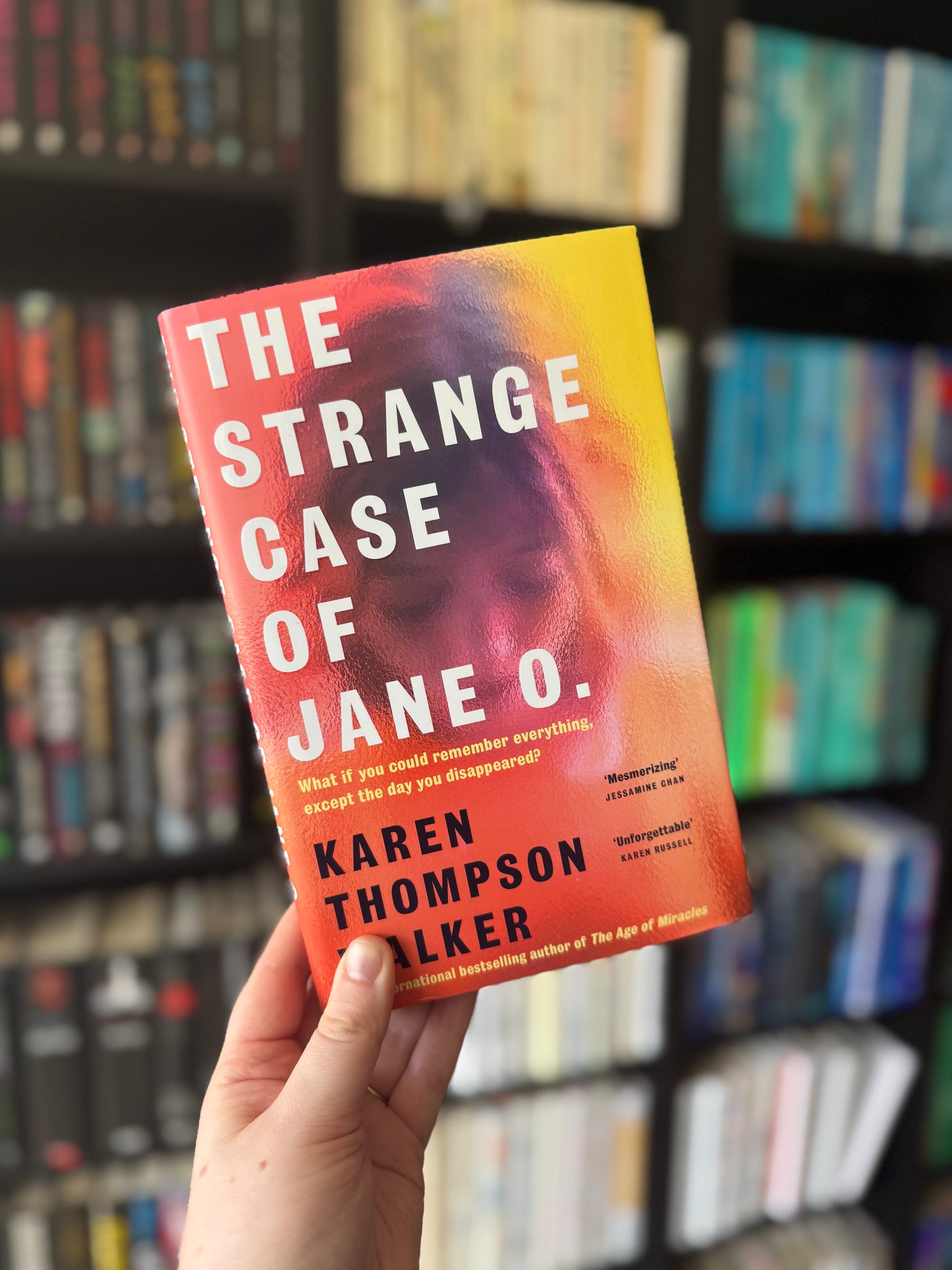 Hand holding "The Strange Case of Jane O." book by Karen Thompson Walker, with a colorful cover in a library setting with blurred bookshelves.