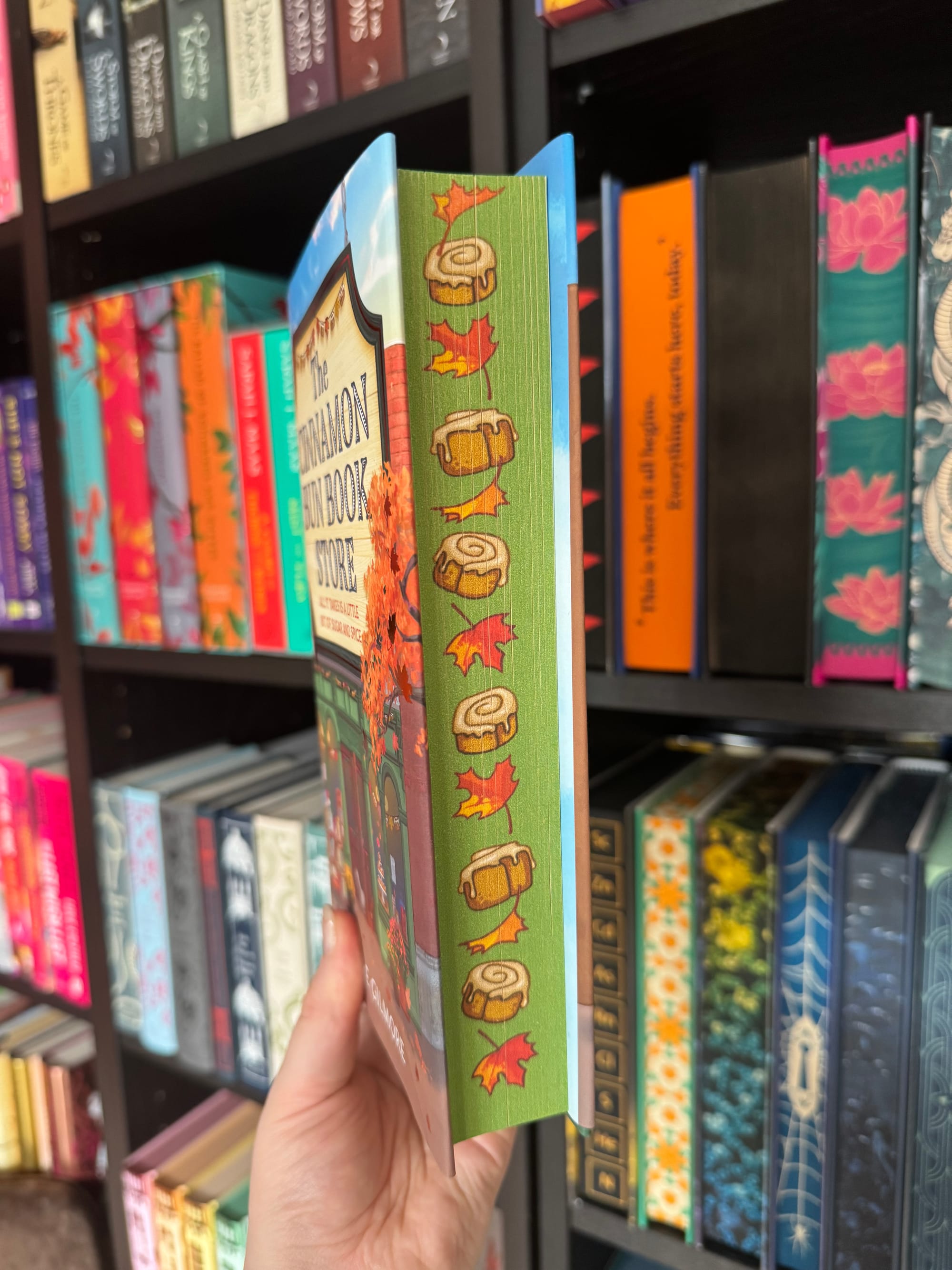 A hand holds a colorful book with cinnamon roll and leaf art on the cover. Shelves filled with brightly patterned books are in the background.