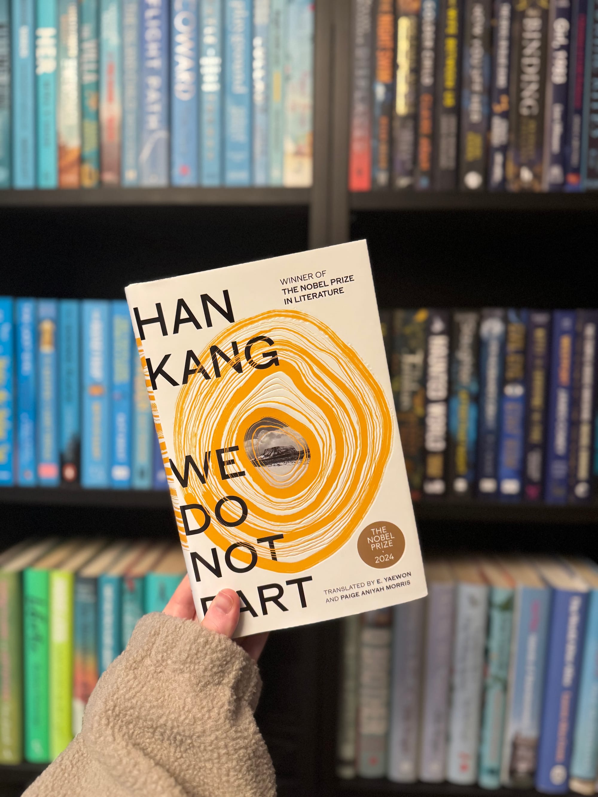 A hand holds the book "We Do Not Part" by Han Kang in front of colorful bookshelves. The cover features bold text and circular patterns.