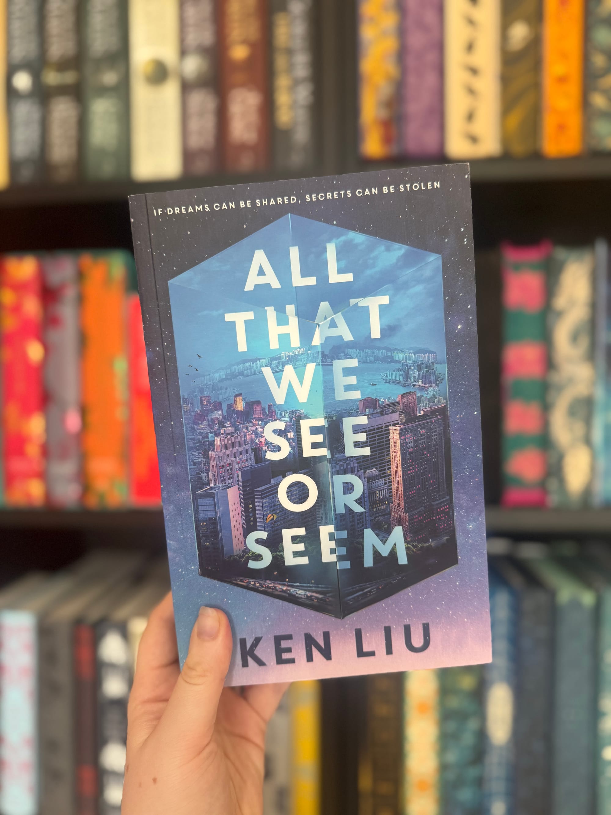 Hand holding a book titled "All That We See or Seem" by Ken Liu, with a cityscape cover. Blurred bookshelf background.