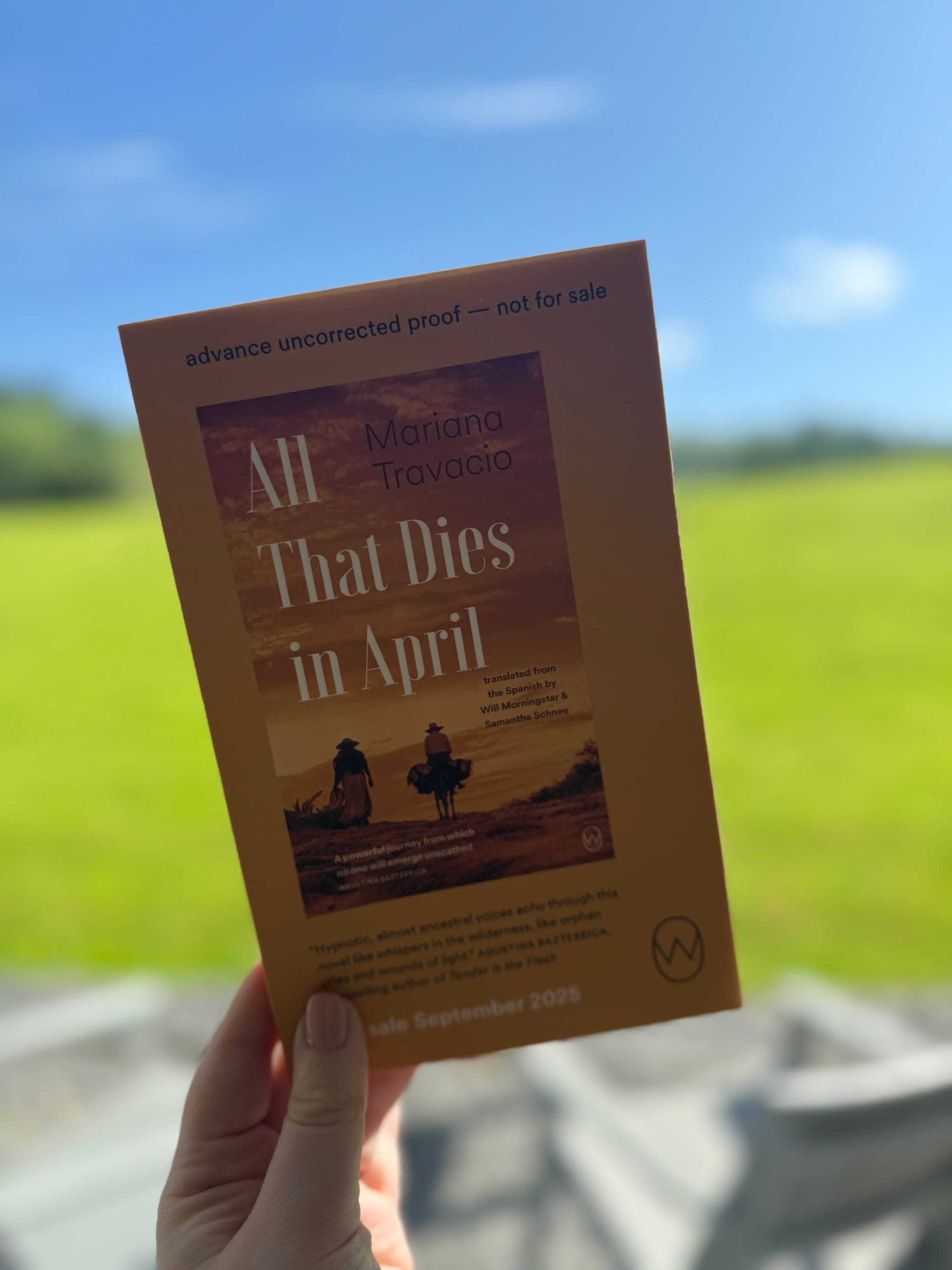 A hand holds a book titled "All That Dies in April" by Mariana Travacio, against a blurry outdoor backdrop. Text notes it's an advance proof.