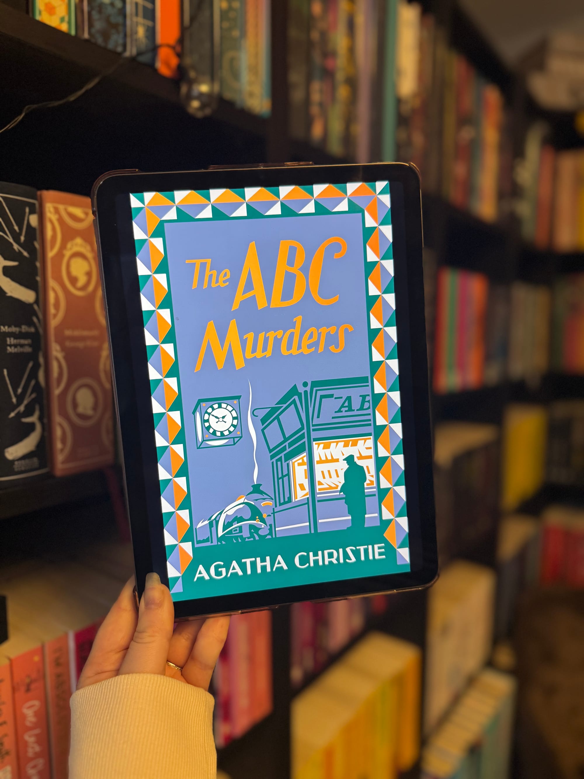 Tablet displaying "The ABC Murders" book cover by Agatha Christie, held in hand. Bookshelves in the background create a cozy ambiance.