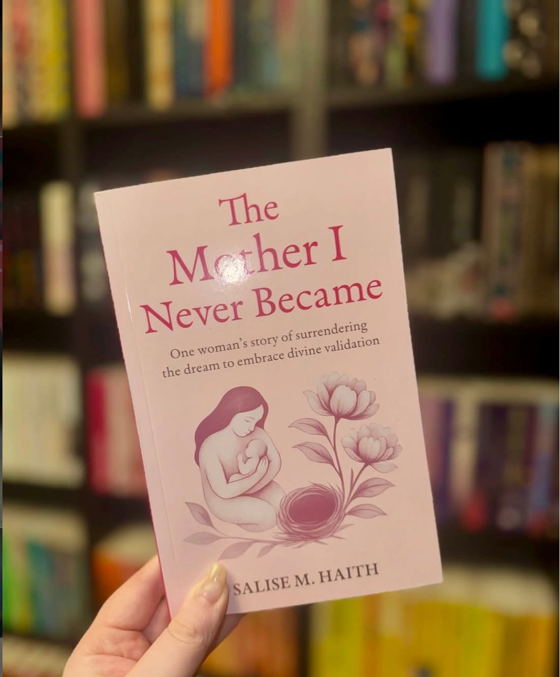 Hand holding a pink book titled "The Mother I Never Became" with an illustration of a mother. Blurred bookshelf in the background.