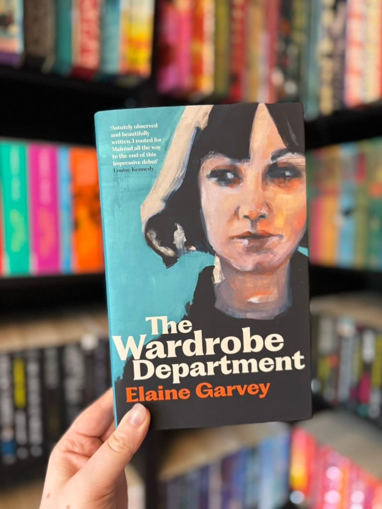 A hand holds a copy of The Wardrobe Department by Elaine Garvey in front of softly blurred rainbow bookshelves. The cover features an expressive painted portrait of a dark-haired woman against a turquoise background, with the title in bold white lettering and the author's name in bright orange. The muted, artistic cover reflects the thoughtful and emotional tone of the story.