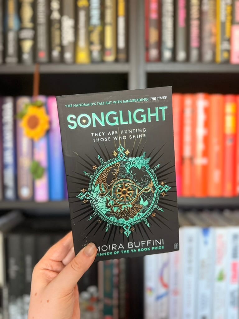 A hand holds a copy of Songlight by Moira Buffini in front of softly blurred rainbow bookshelves. The black cover features intricate teal and gold artwork centred around a compass, with two mirrored figures surrounded by decorative fantasy-inspired details. The tagline, "They are hunting those who shine," appears beneath the title, hinting at the story's dystopian fantasy setting.
