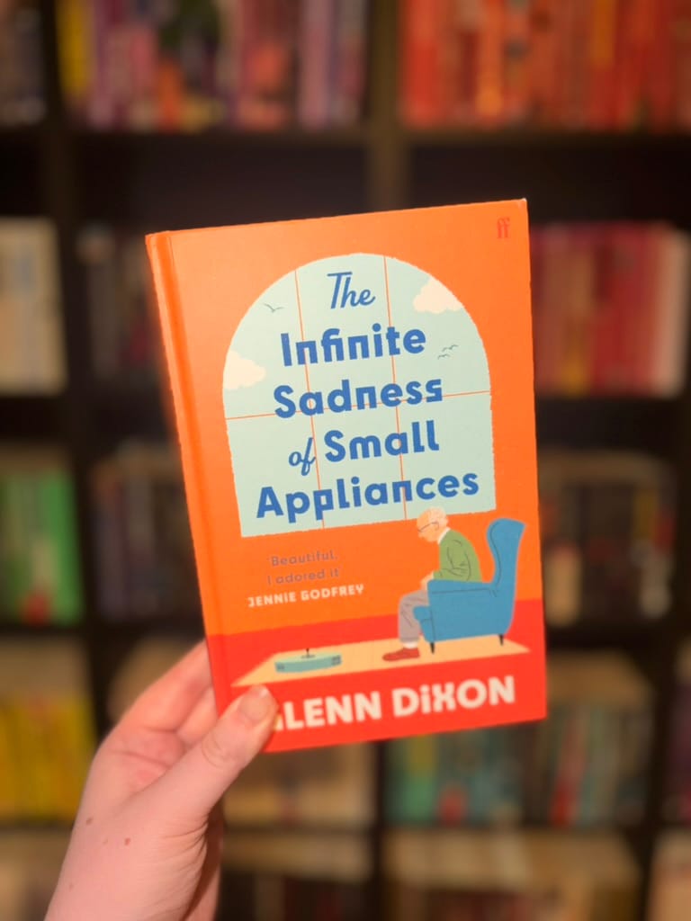 A hand holds a copy of The Infinite Sadness of Small Appliances by Alann Dixon in front of softly blurred rainbow bookshelves. The bright orange cover features an illustration of an elderly man sitting in a blue armchair beside a small robotic vacuum, framed by a large arched window. The warm, minimalist artwork contrasts with the book's poignant title, hinting at a thoughtful and quietly emotional story.