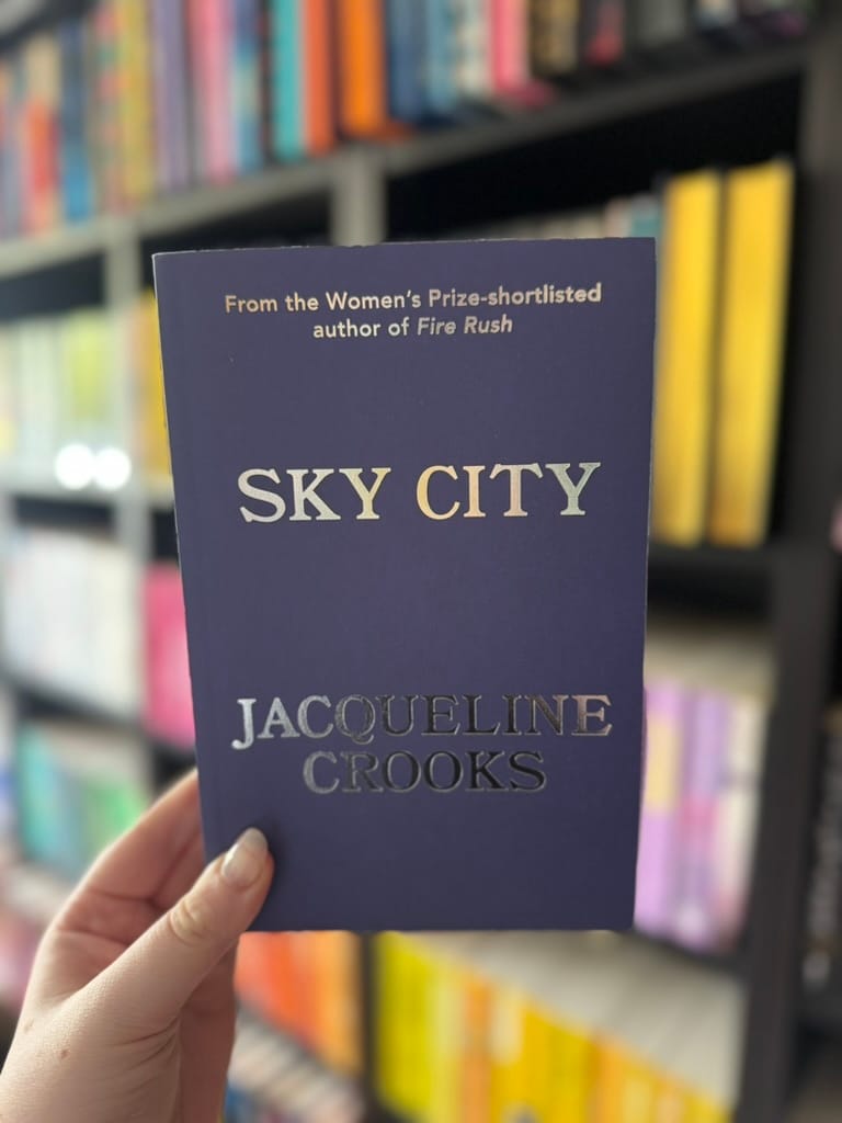 A hand holds the proof copy of Sky City by Jacqueline Crooks in front of softly blurred rainbow bookshelves. The minimalist cover is a deep indigo blue with the title in large white lettering and the author's name in reflective silver foil. Above the title is the text, "From the Women's Prize-shortlisted author of Fire Rush," giving the proof edition a clean and elegant design.