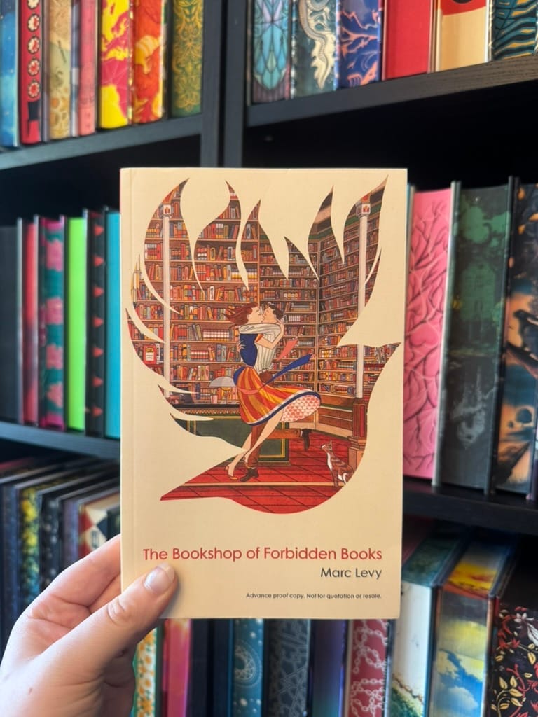 Hand holds The Bookshop of Forbidden Books by Marc Levy in front of colorful bookshelves; cover shows a couple kissing in a library.