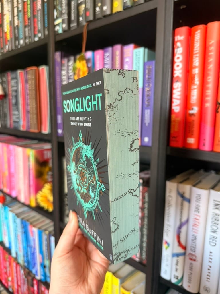 A hand holds Songlight by Moira Buffini at an angle to showcase the sprayed page edges. The edges feature a pale green map design with illustrated coastlines and place names, complementing the teal and black cover. The special edition detailing is photographed against colourful, softly blurred bookshelves, highlighting the decorative sprayed edges.